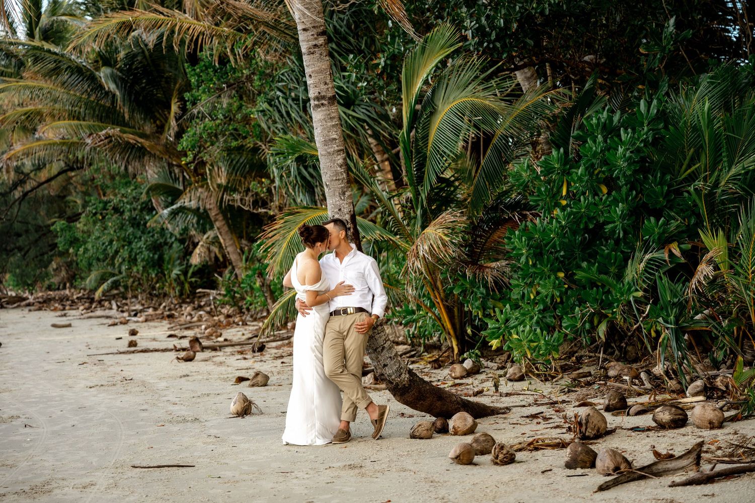 Newlyweds share a romantic kiss surrounded by coconuts on a lush tropical beach at Four Mile Beach, Port Douglas.
