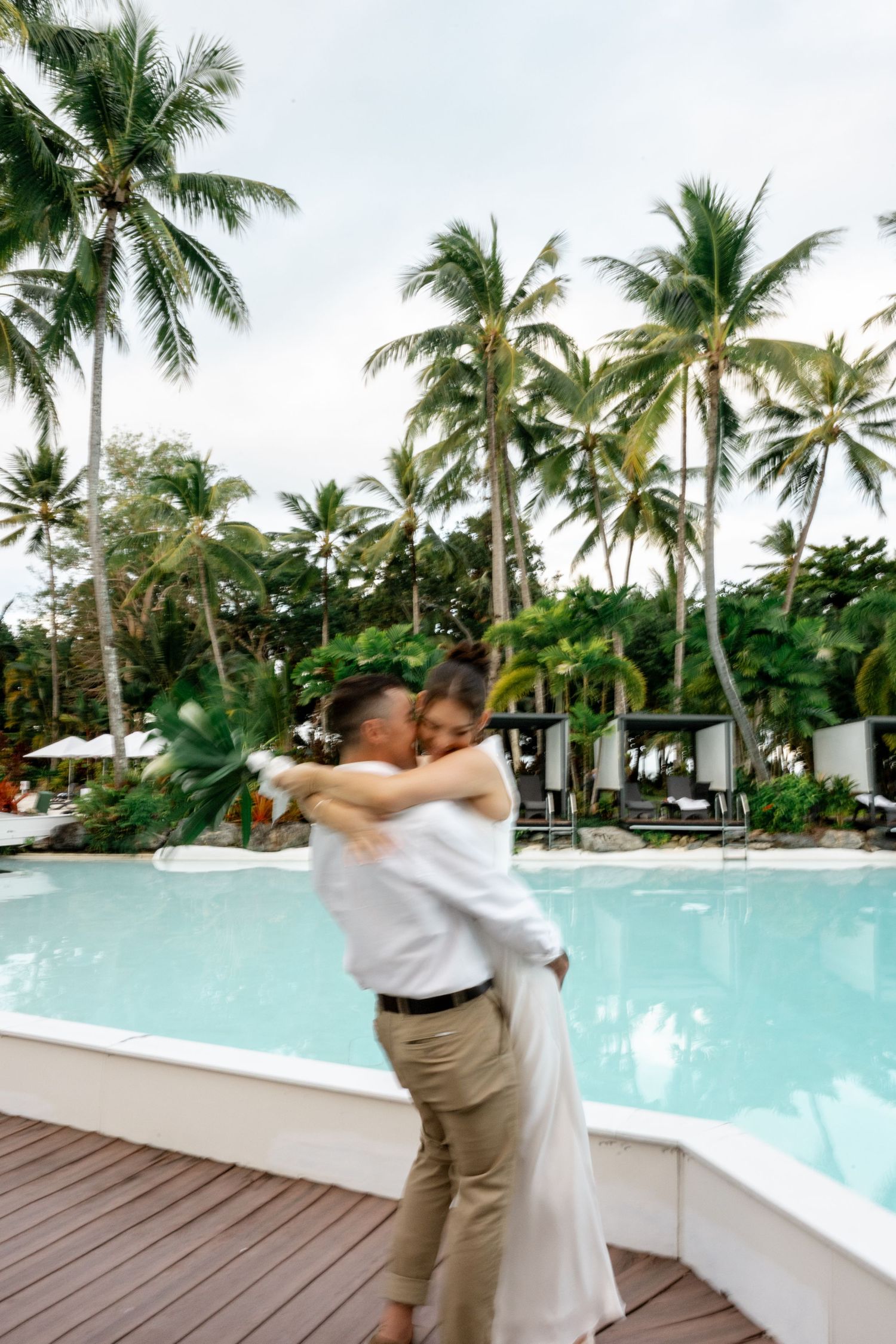 Couple embracing and spinning joyfully by a tropical resort pool surrounded by tall palm trees at Sheraton Grand Mirage Port Douglas.
