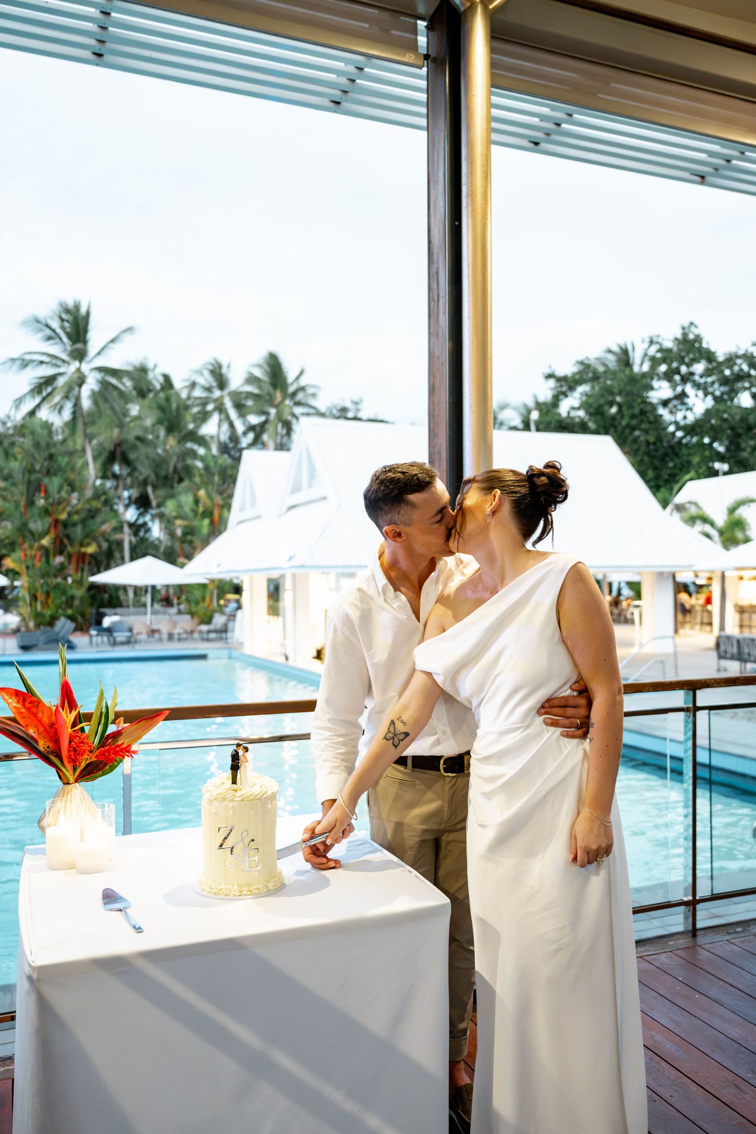 Newlyweds kissing while cutting their white wedding cake at a tropical poolside reception venue, Sheraton Grand Mirage Port Douglas.