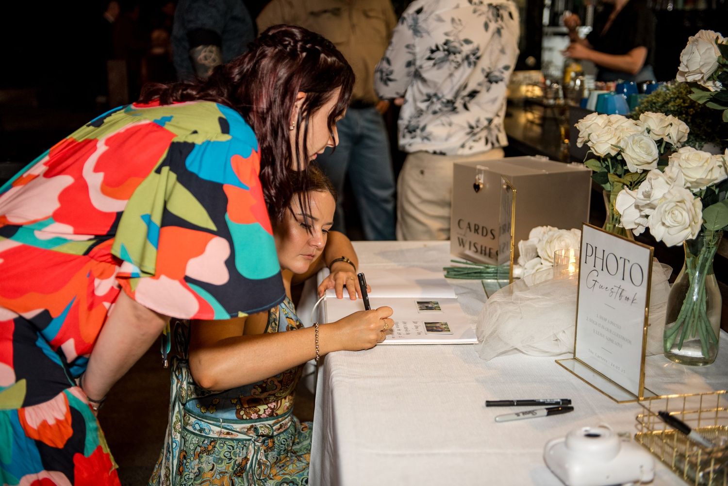 Two female wedding guests lean over to sign the guestbook at a floral decorated reception table.