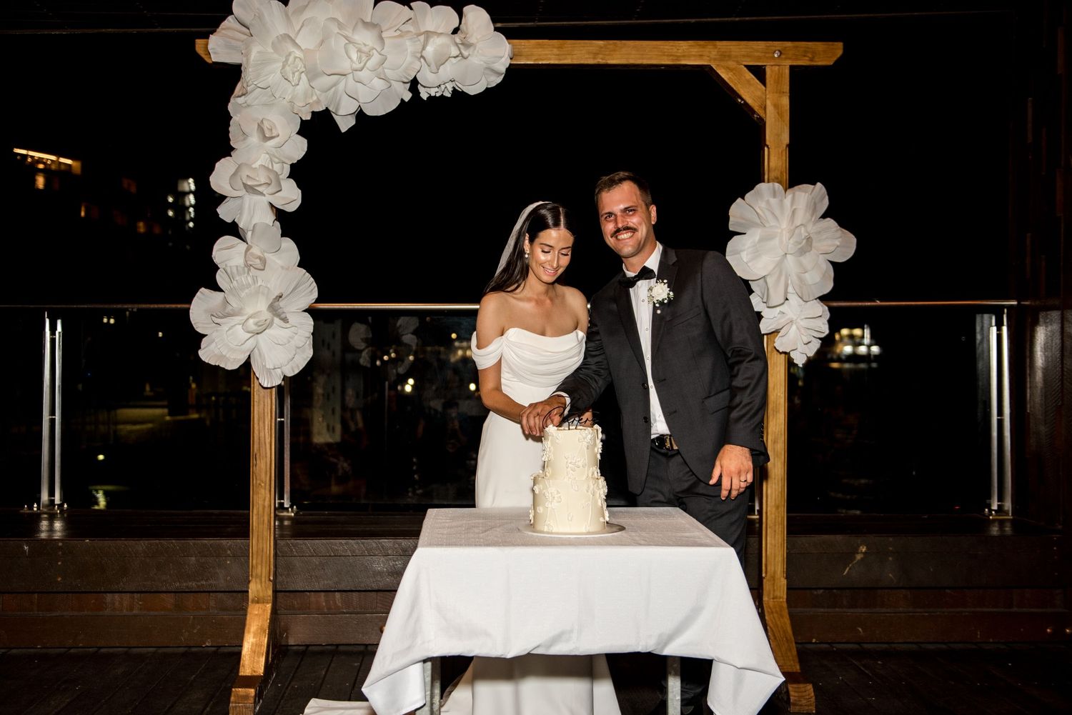 Bride and groom smile while cutting their white wedding cake under a wooden arch with balloon decorations at night.