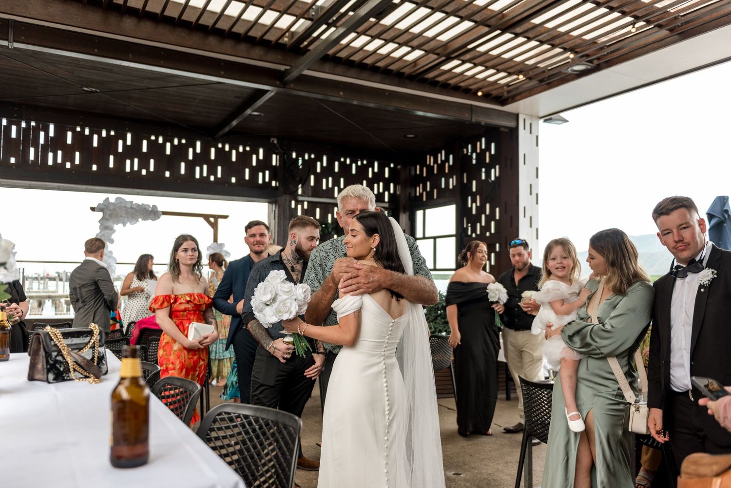 Bride in white veil hugging guest warmly while surrounded by wedding guests at reception.