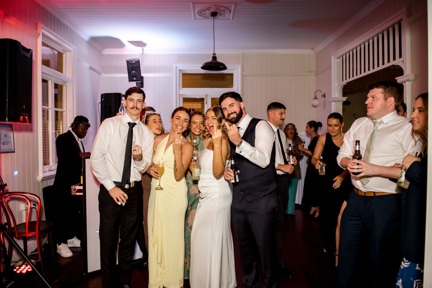 Bride and groom posing and smiling with wedding guests holding drinks at an indoor reception celebration.