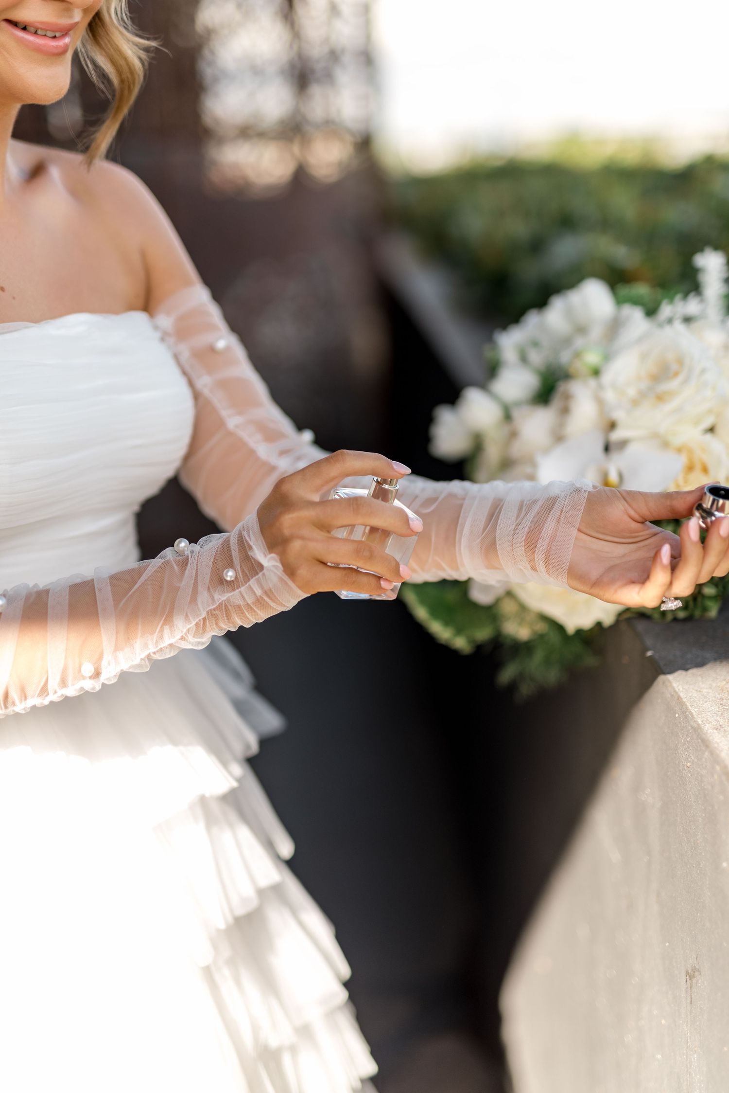 Bride in ruffled wedding gown with sheer gloves applying perfume while holding white floral bouquet.