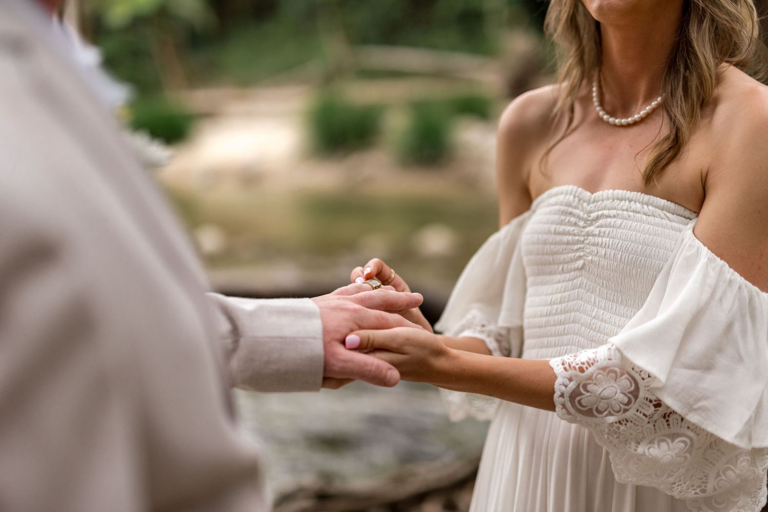 Bride and groom hold hands tenderly during heartfelt outdoor wedding ceremony by forest river.