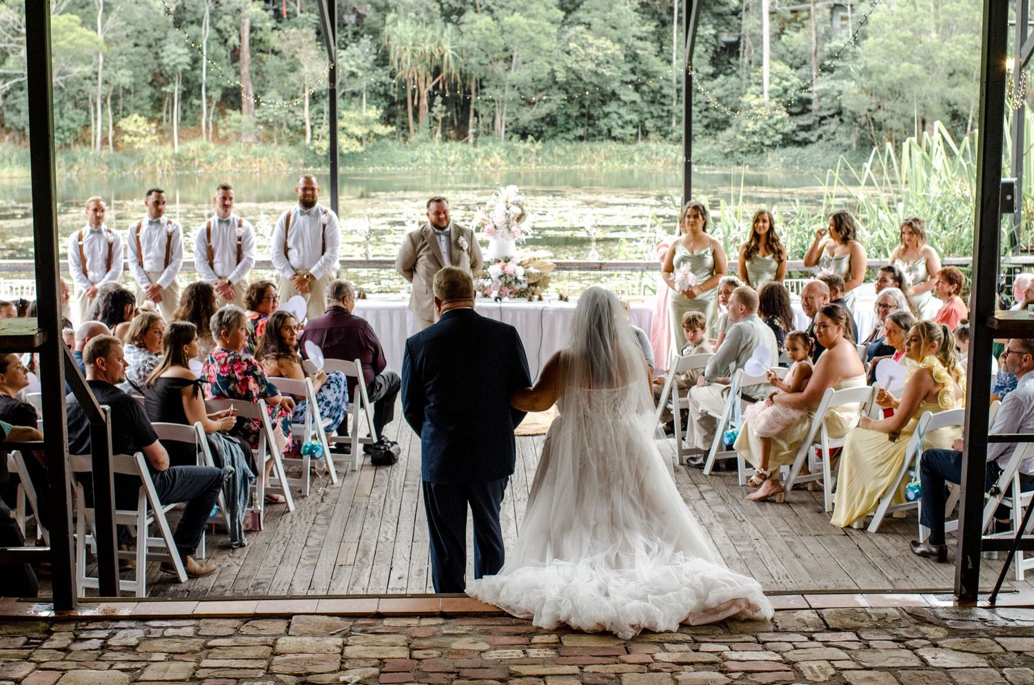 Bride and escort walk toward altar at outdoor wedding ceremony by a scenic pond.