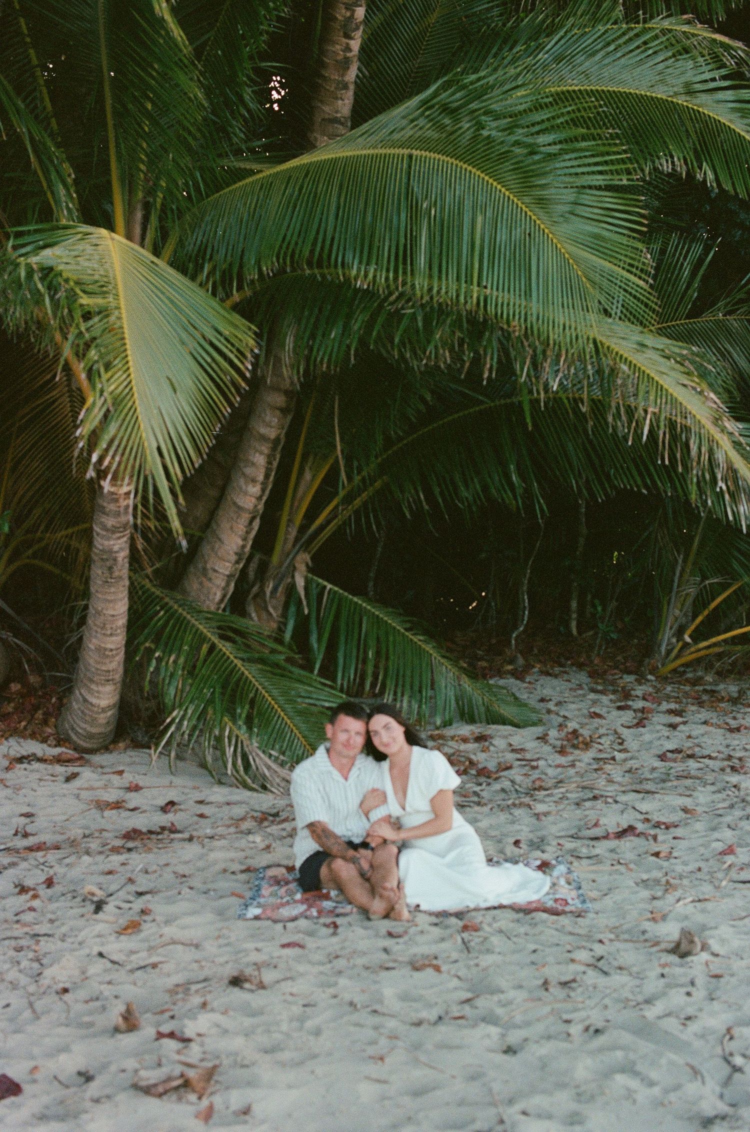 Happy engaged couple sitting together on sandy tropical beach surrounded by lush palm trees.