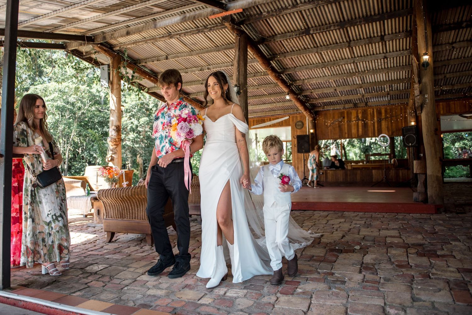 Bride in white gown walks with young ring bearer and guest under rustic wooden pavilion.