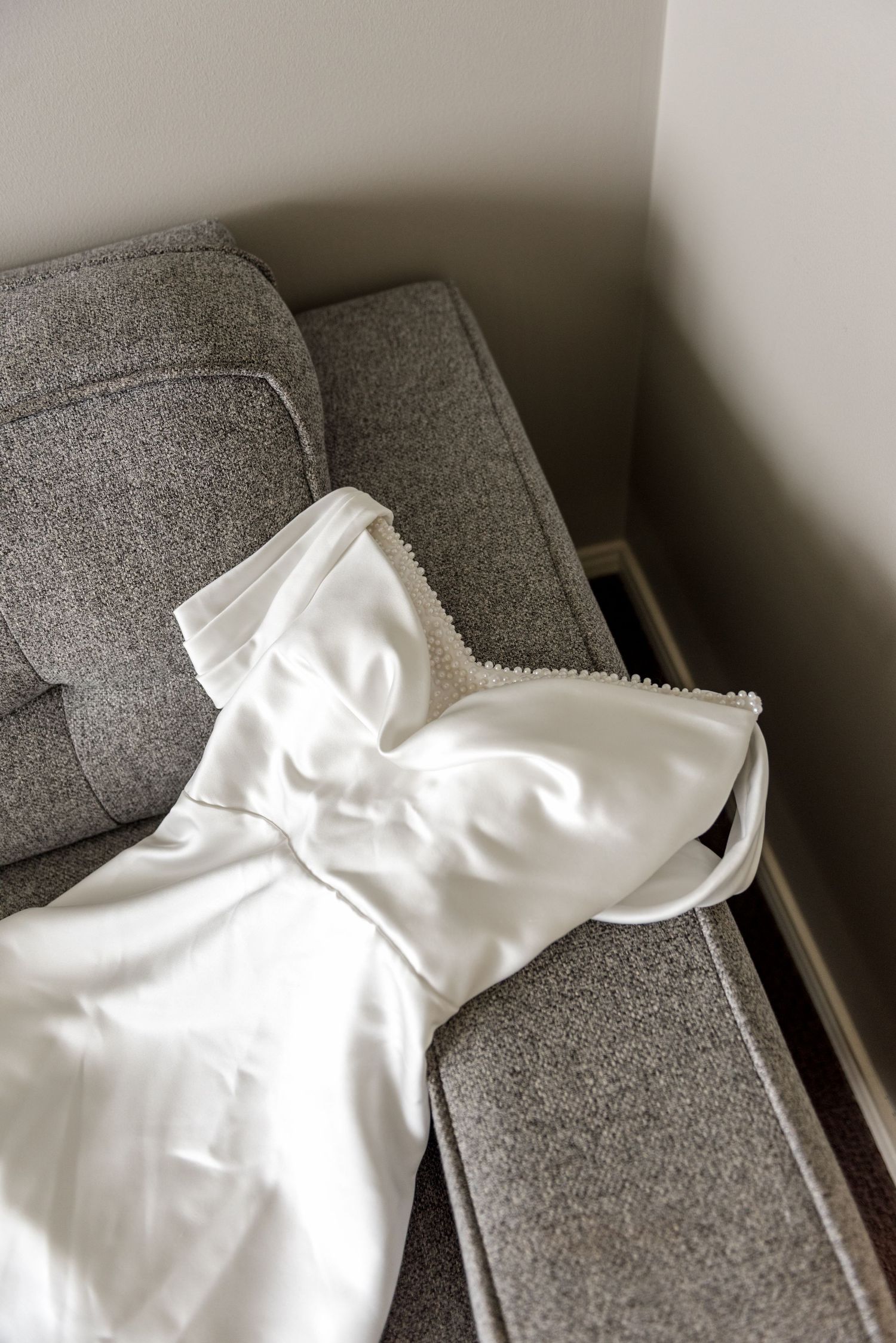 Elegant white strapless wedding dress with pearl trim detail laid on grey sofa before ceremony.