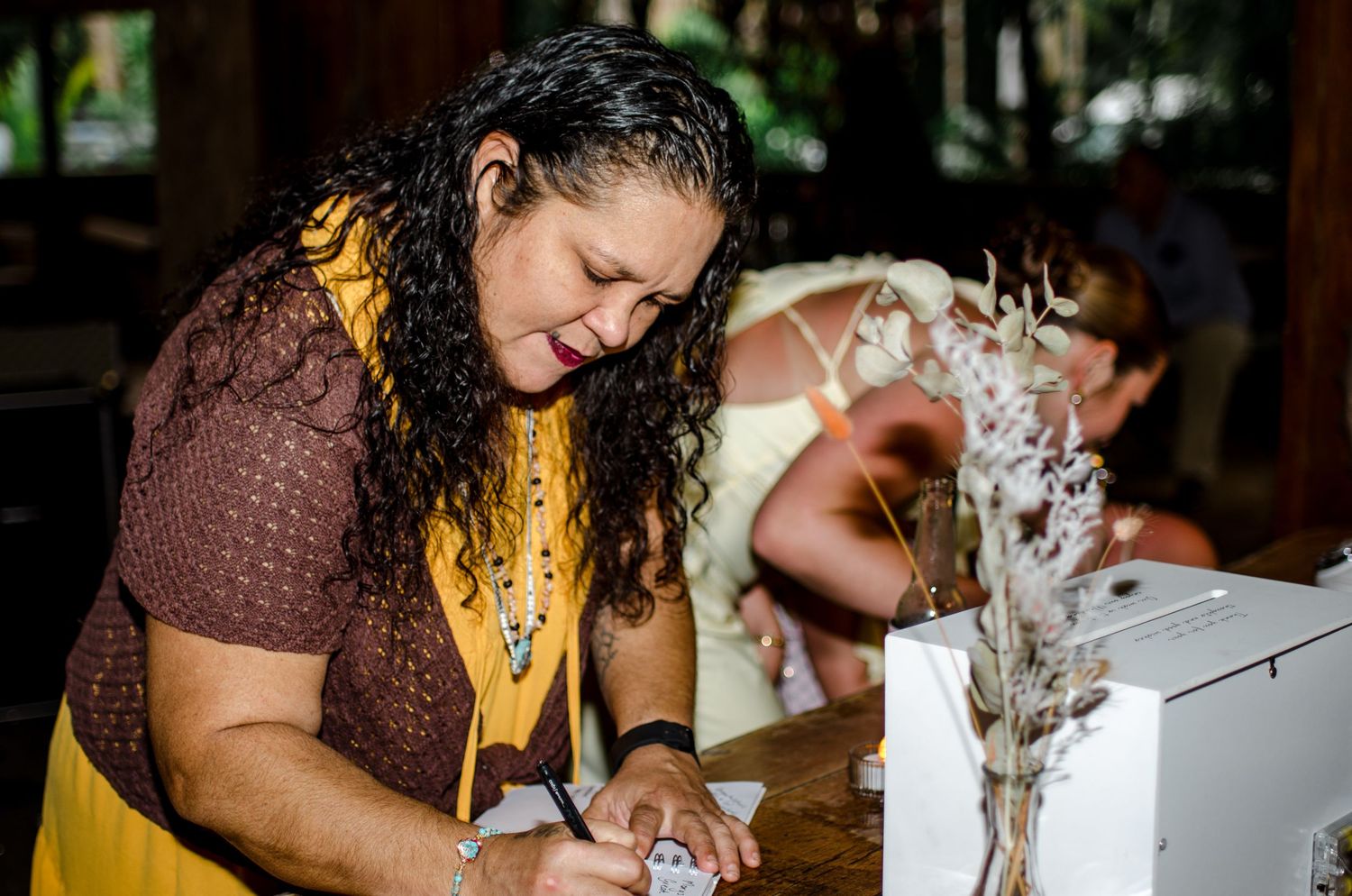 Woman in yellow and brown outfit signs a document at a table decorated with dried flowers.