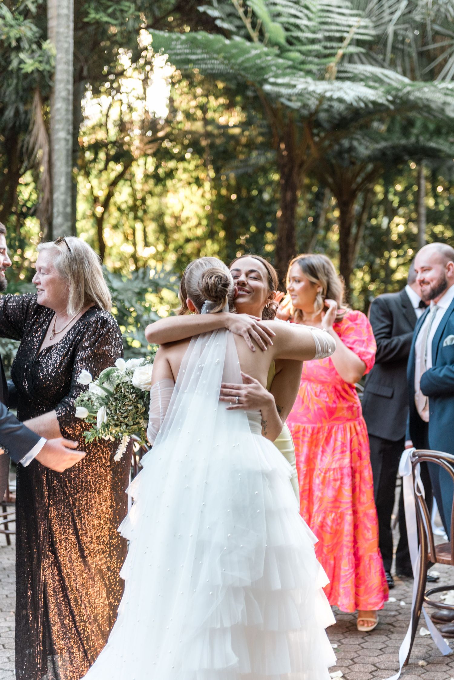 Bride in tiered white wedding gown receives a warm hug from a guest during outdoor wedding ceremony reception.