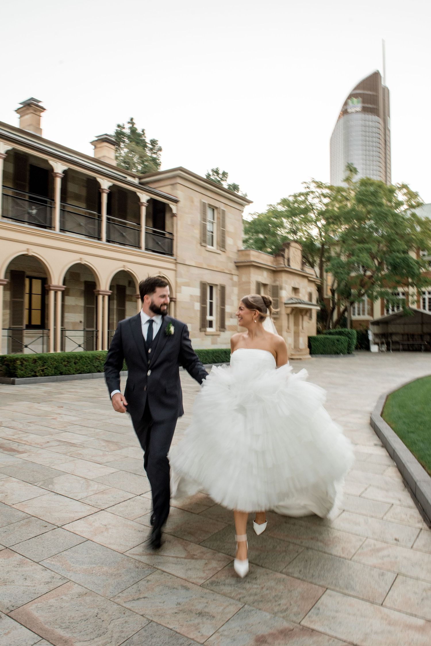 Bride in ruffled strapless gown and groom in dark suit walk hand-in-hand outside historic stone building.
