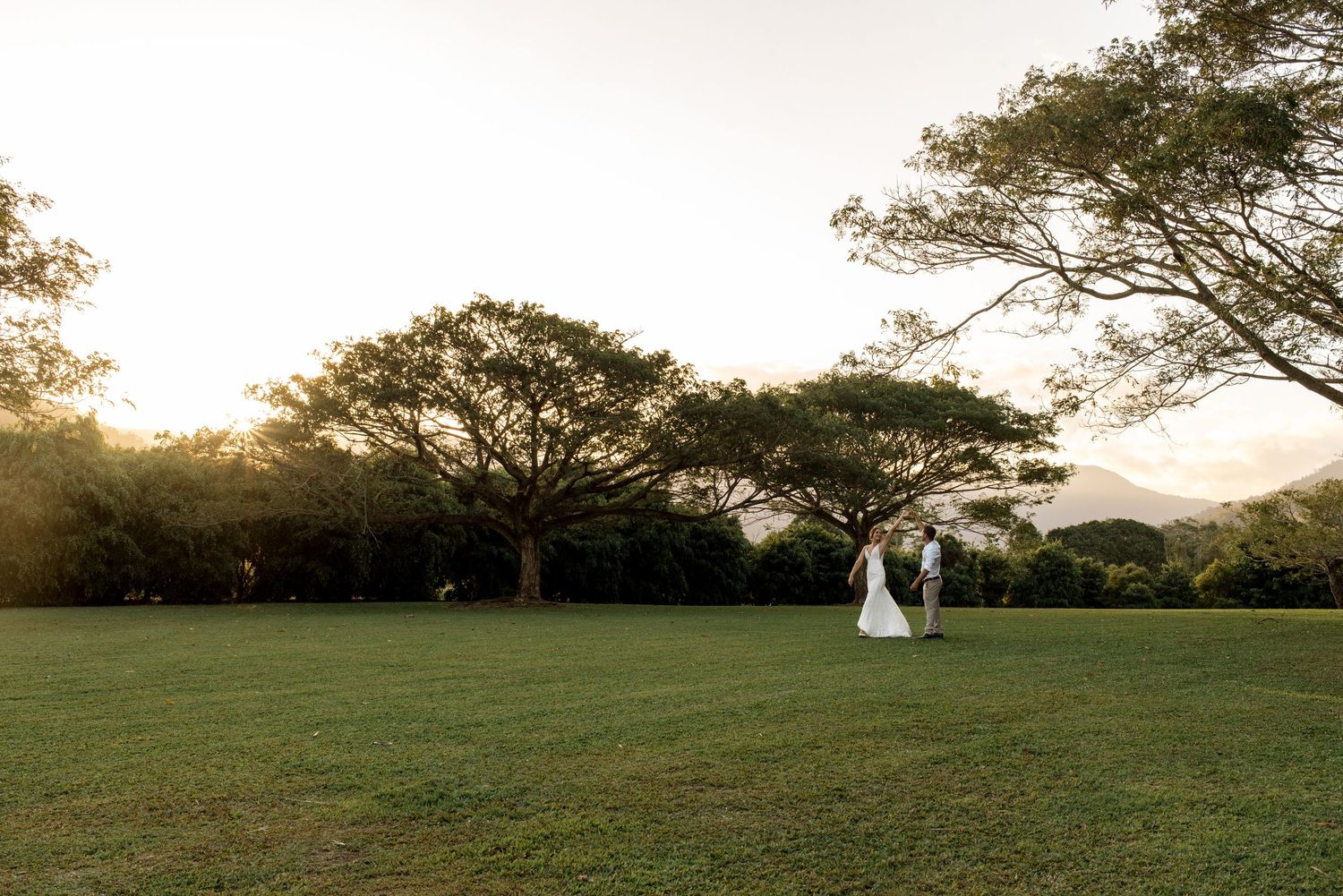 Bride and groom walking across expansive green lawn beneath large trees at golden sunset.