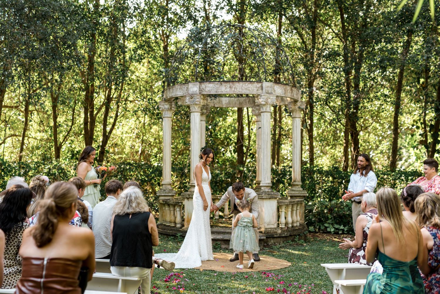 Intimate outdoor wedding ceremony at stone gazebo with small gathering of guests seated on white benches.