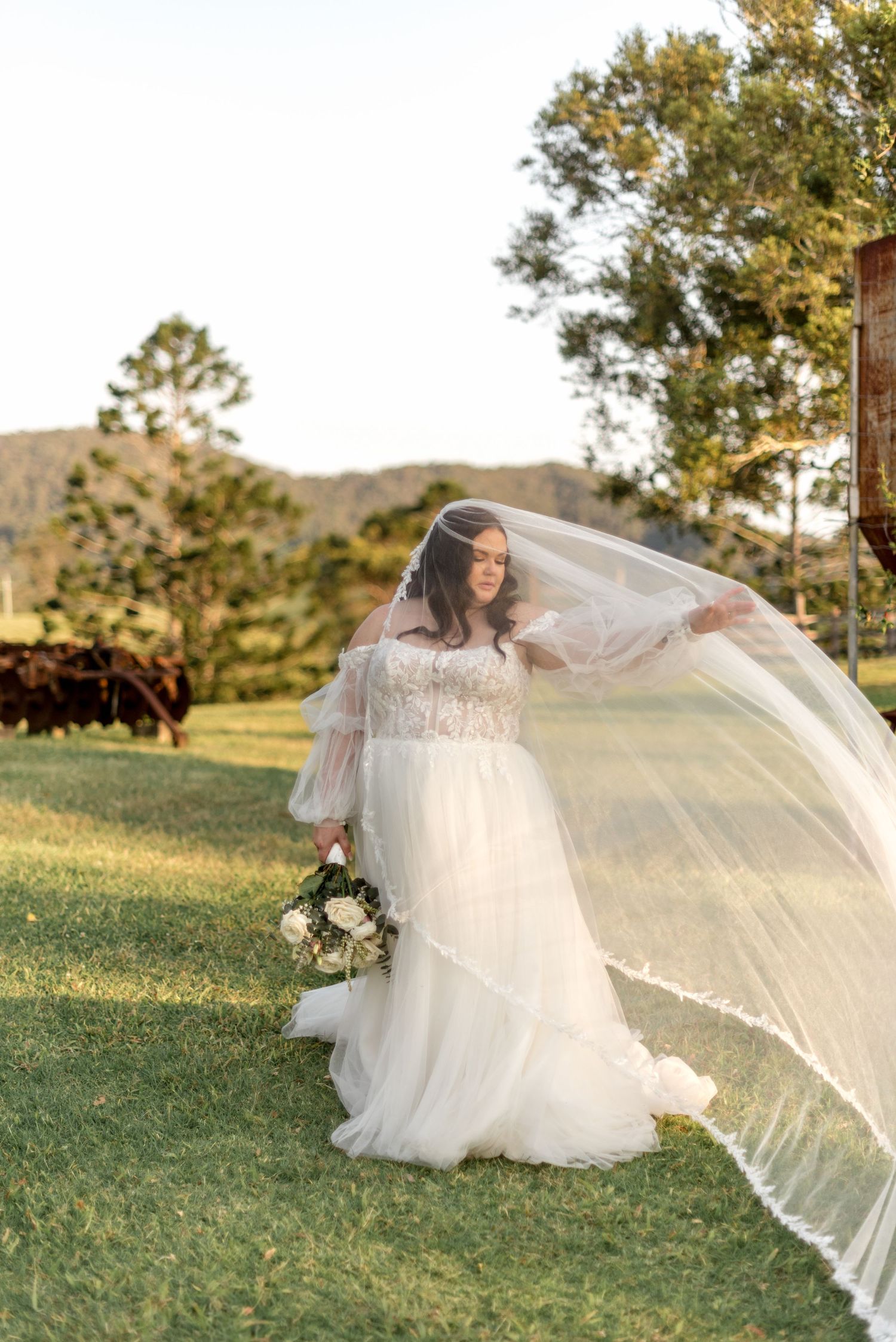 Bride in white gown with veil flowing in breeze holding bouquet in sunlit field.