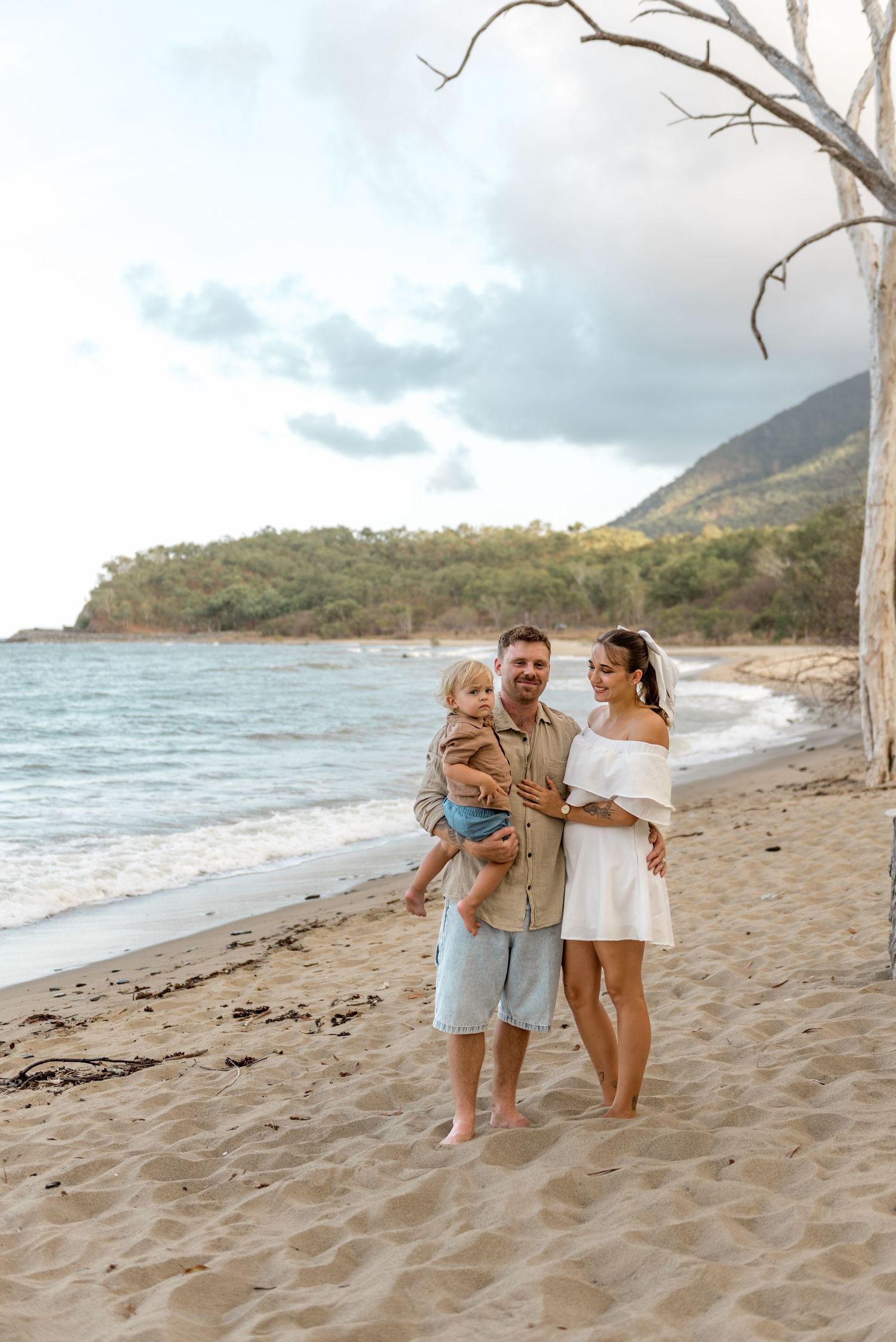Smiling family with young child posing on sunny beach with turquoise ocean and hills backdrop.