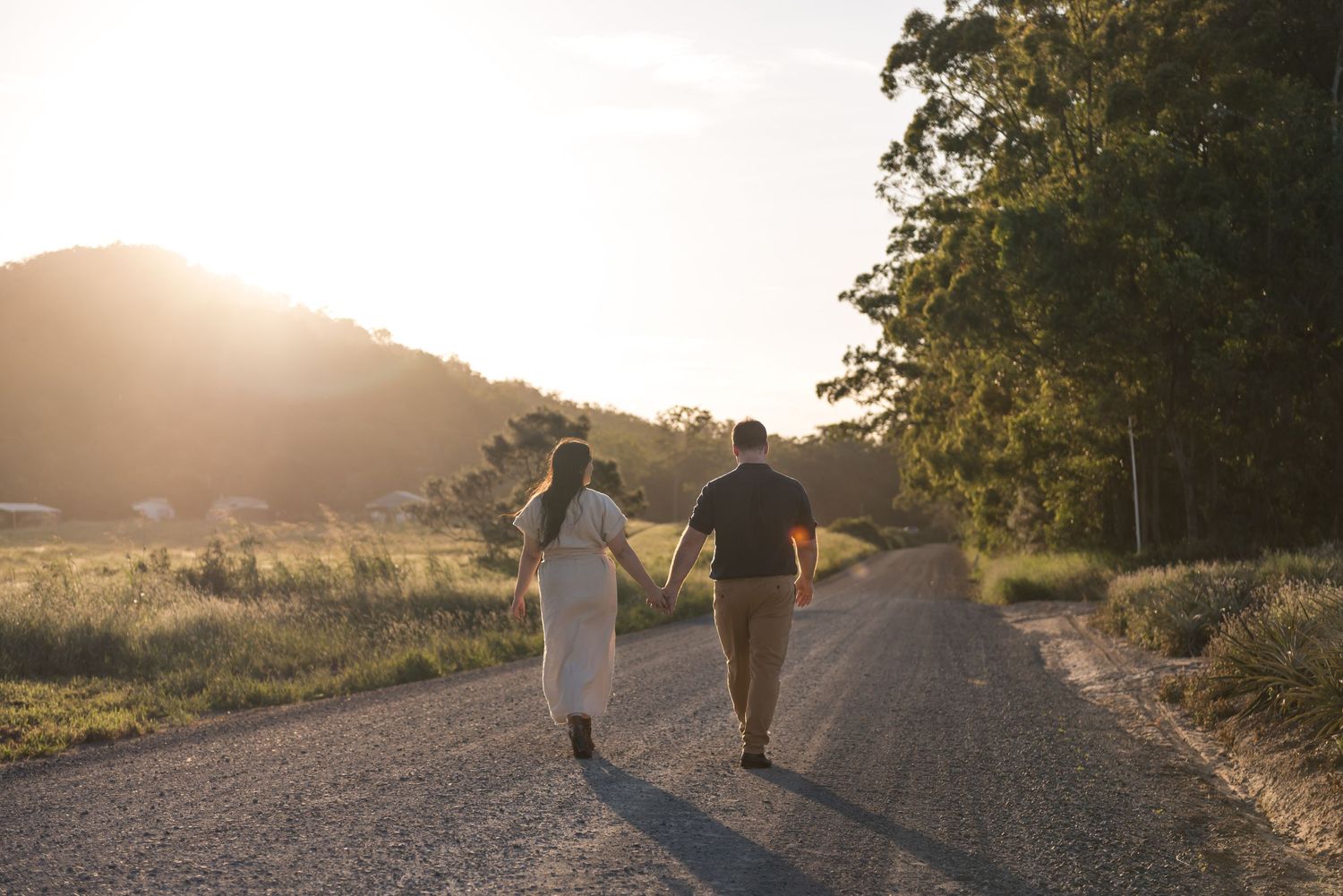 Couple walking hand in hand down scenic gravel road toward golden sunset hills.