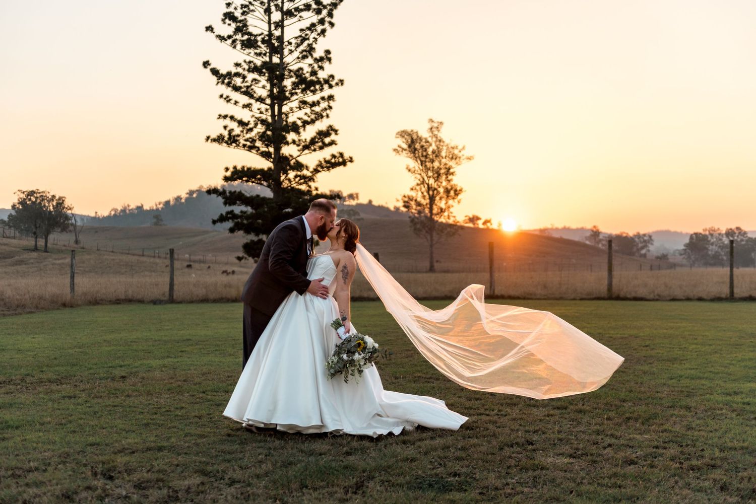 Bride and groom sharing a kiss with veil flowing in the breeze at golden sunset outdoors.
