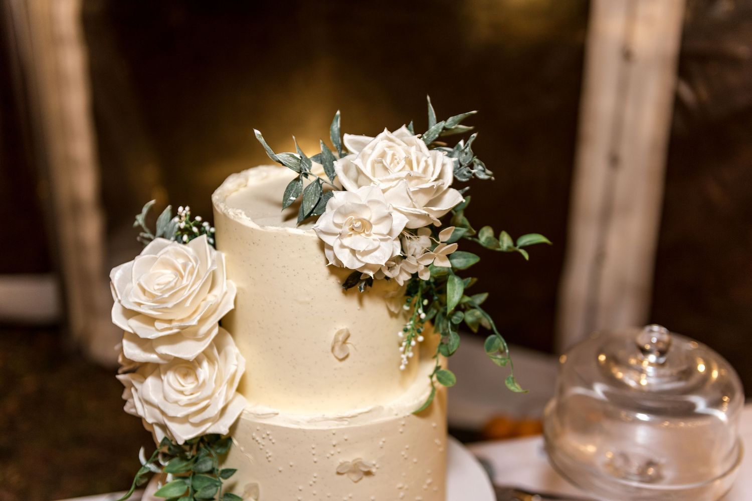 Elegant two-tier white wedding cake adorned with large sugar roses and greenery on a rustic wooden table.
