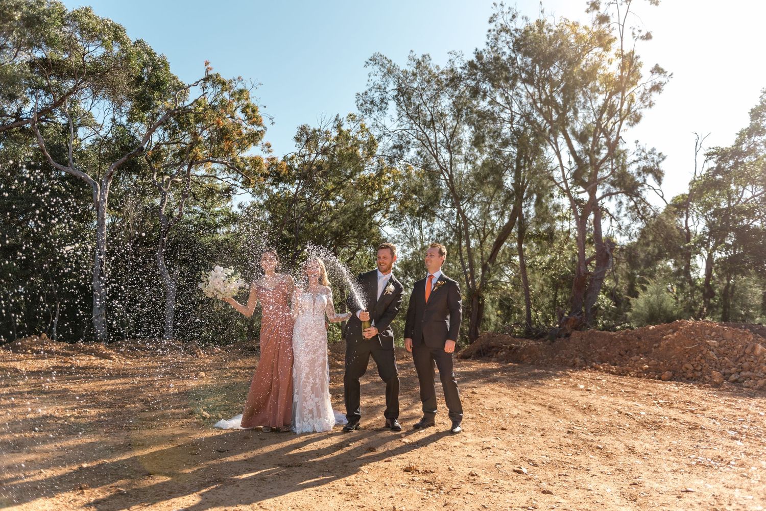 Bride and groom celebrating with champagne spray outdoors with groomsman in sunny forest setting.
