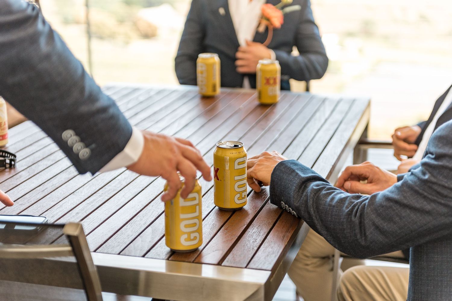 Groomsmen grabbing cold beer cans from a wooden outdoor table during wedding preparations.