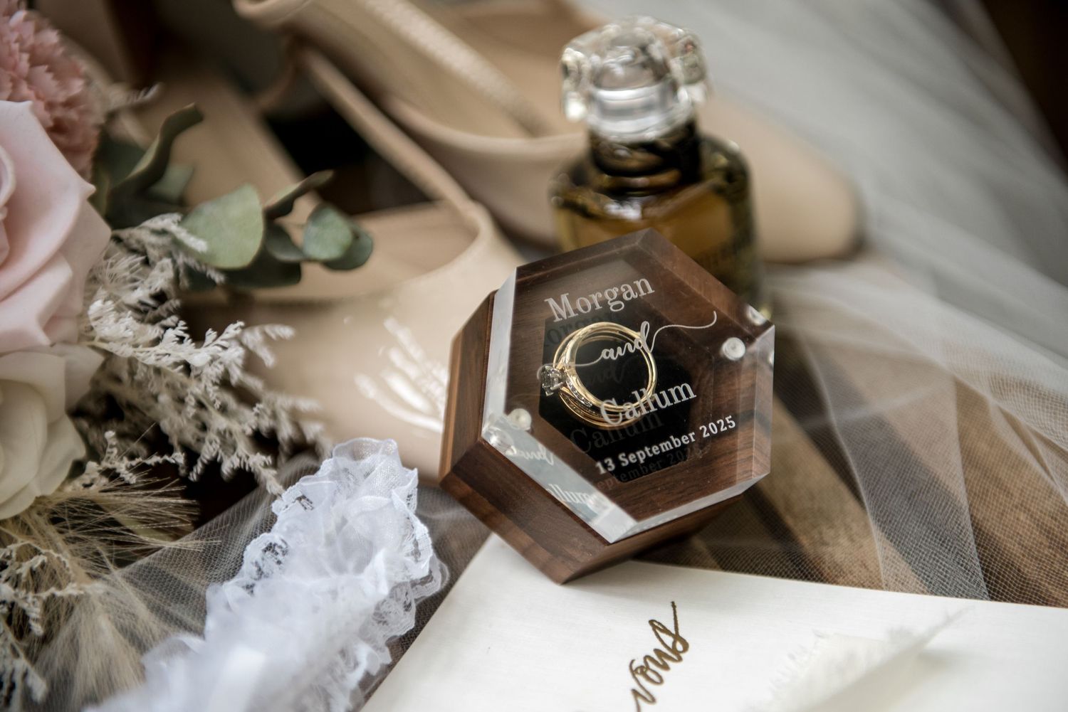Wedding rings displayed in a hexagonal wooden ring box engraved with Morgan and Callum beside bridal accessories.