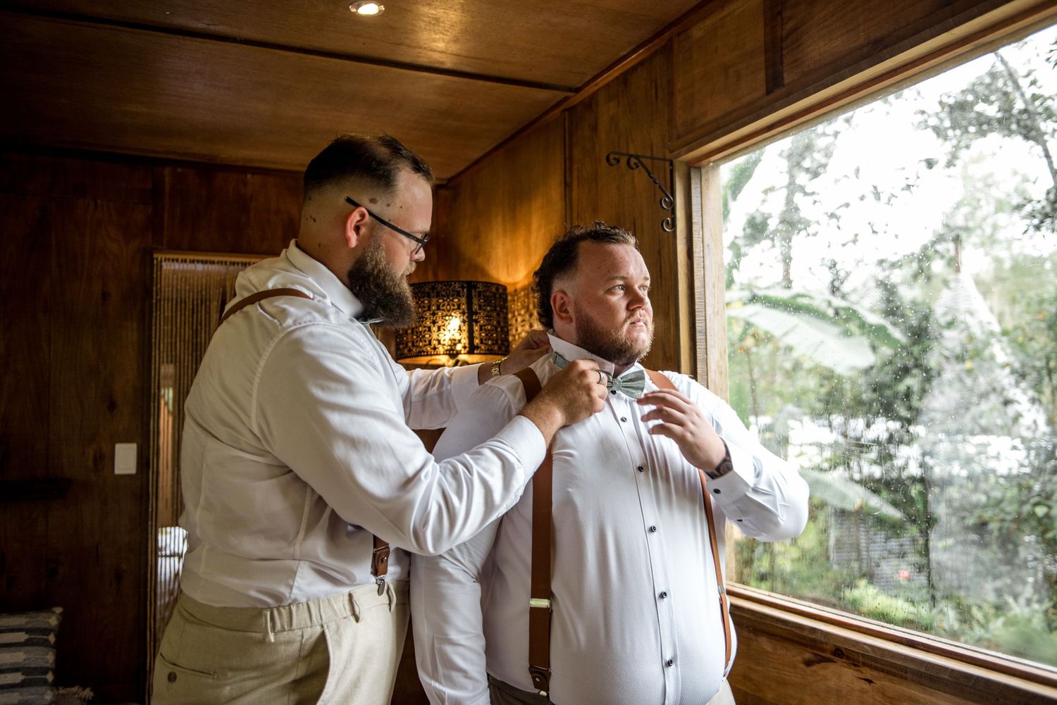 Groomsman helping groom adjust his bow tie while getting ready in a warmly lit wooden cabin.