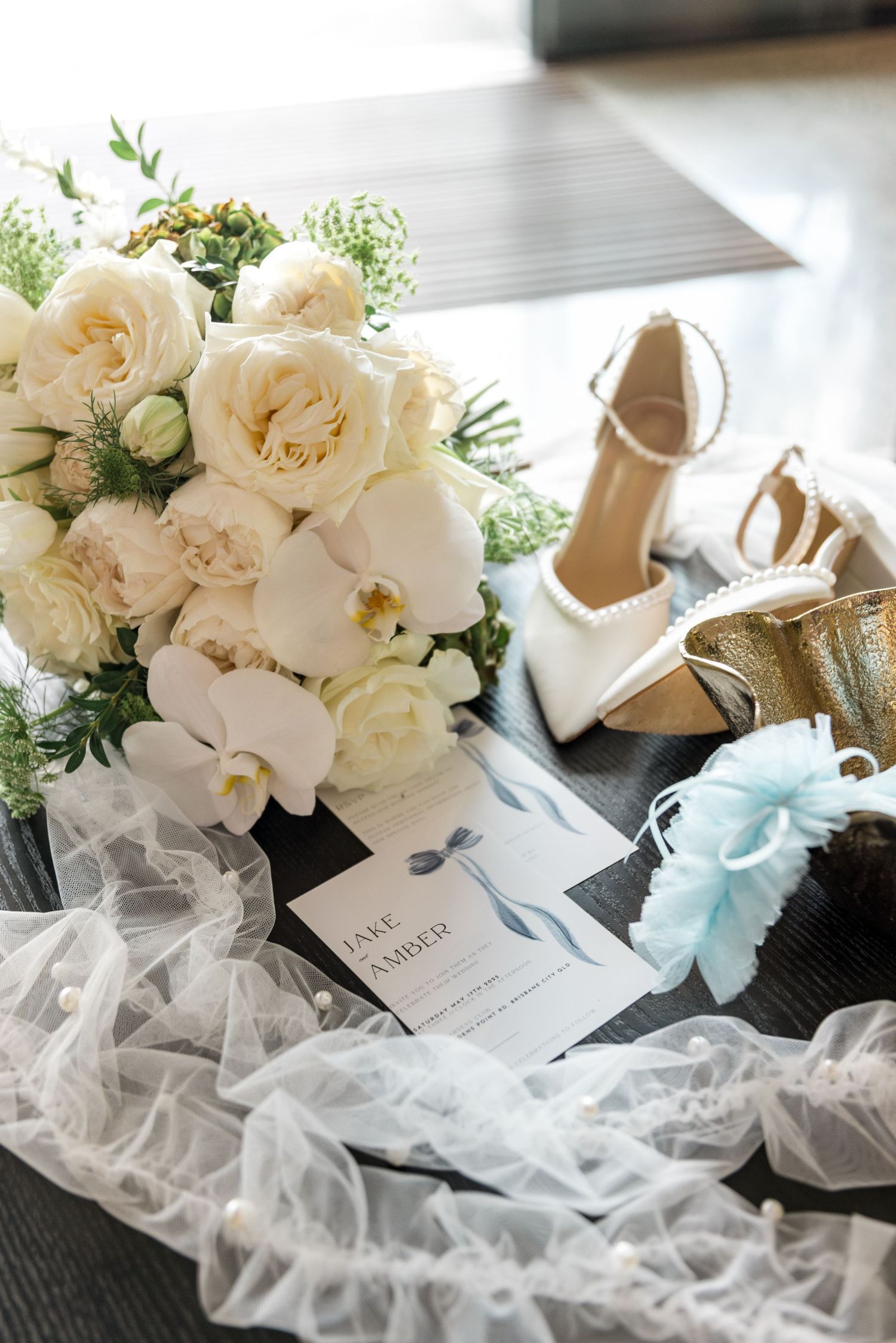 Bridal flat lay with white rose bouquet, wedding invitation, heels, garter, and veil.