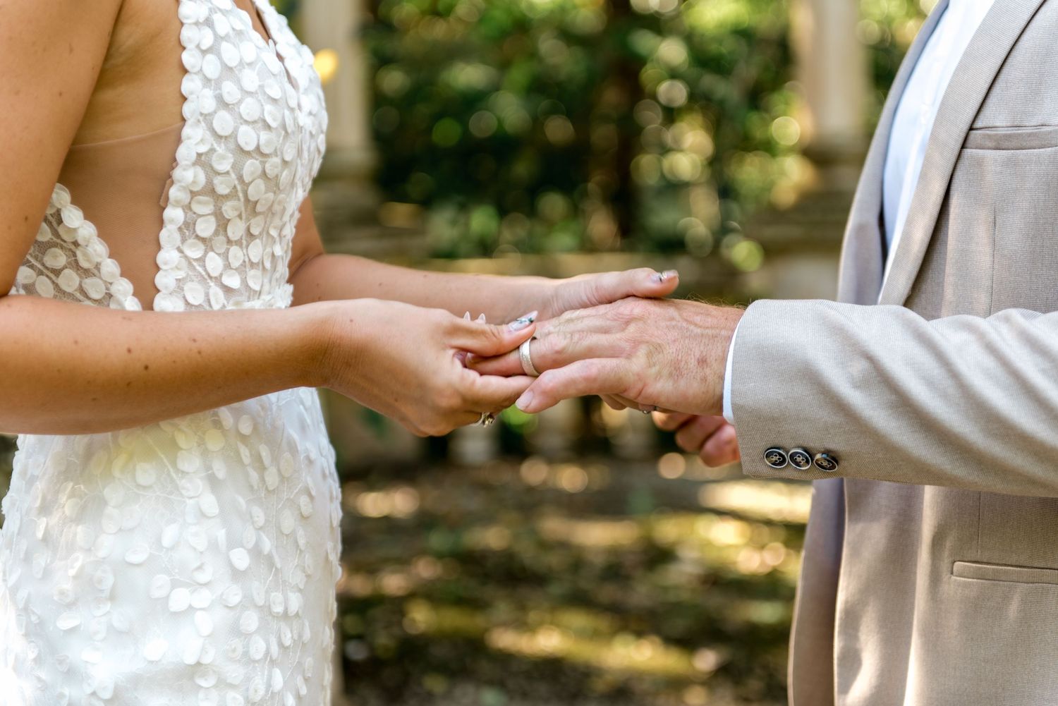 Close-up of groom placing wedding ring on brides finger during outdoor ceremony, bride in lace dress.