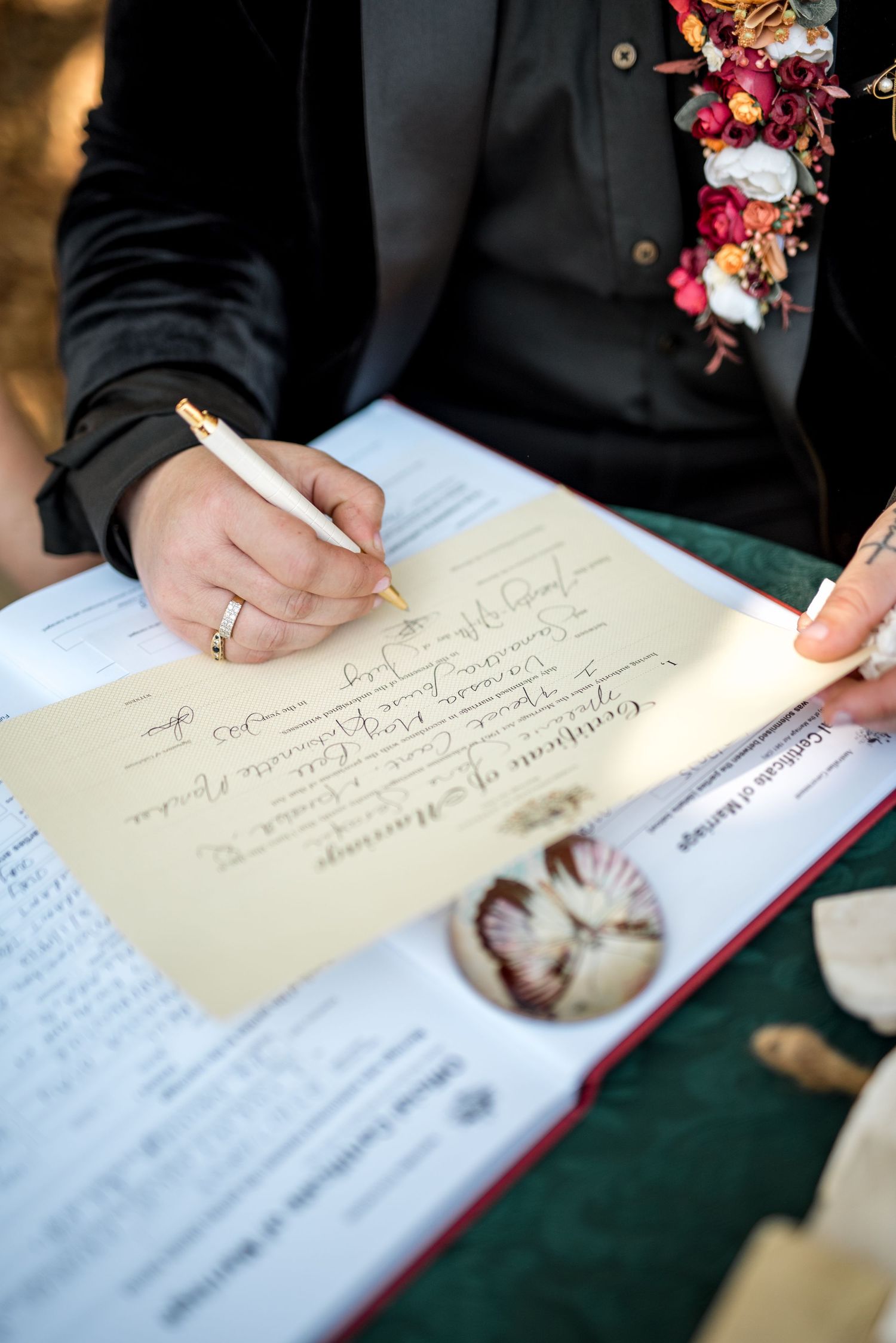 Groom in black suit signs wedding certificate with gold pen at green velvet table.