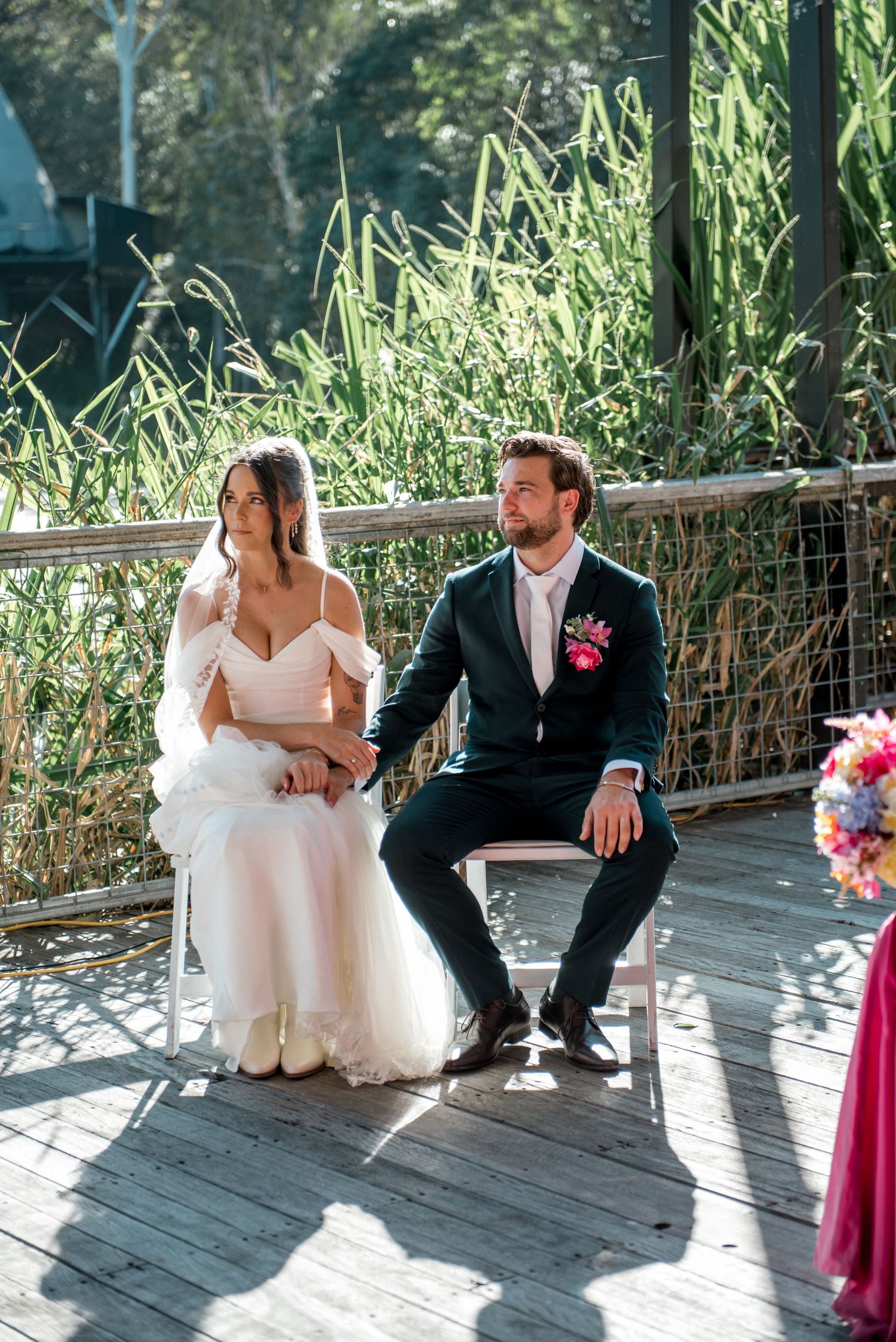 Bride in white gown and groom in dark suit sit together on wooden deck during wedding ceremony.