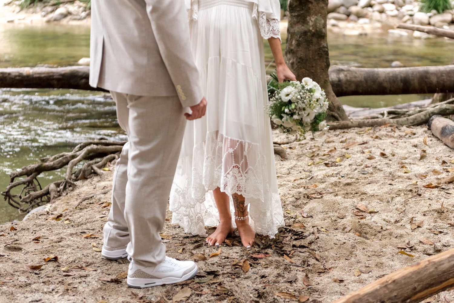 Close-up of bride holding white bouquet in lace wedding dress beside groom in sandy forest.