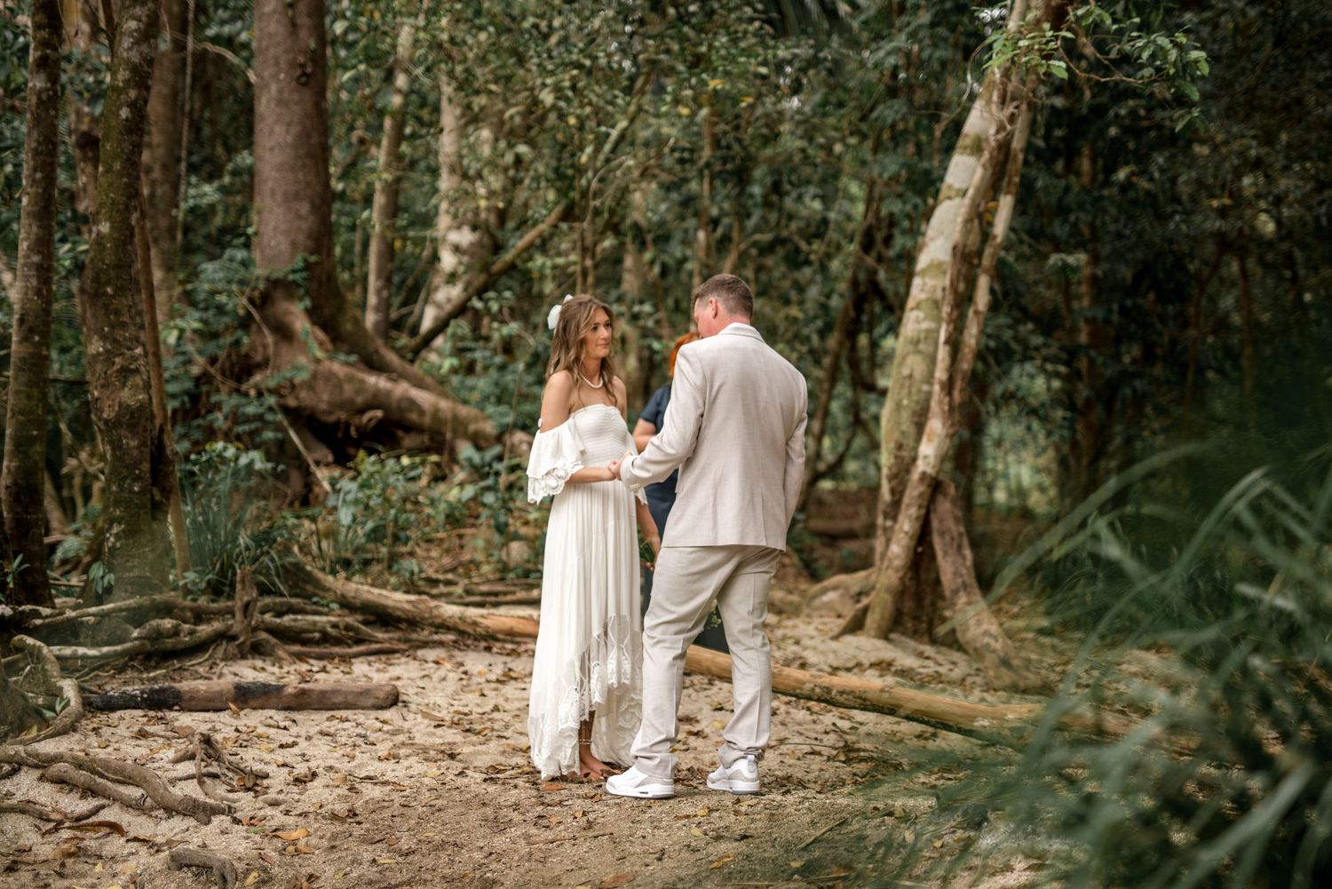 Bride in white off-shoulder gown and groom in beige suit exchange vows in forest.