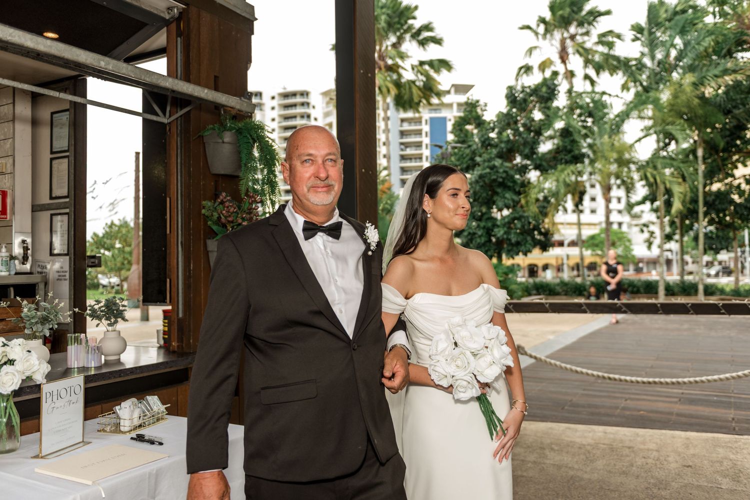 Bride in off-shoulder gown walks arm-in-arm with bald man in tuxedo outdoors.