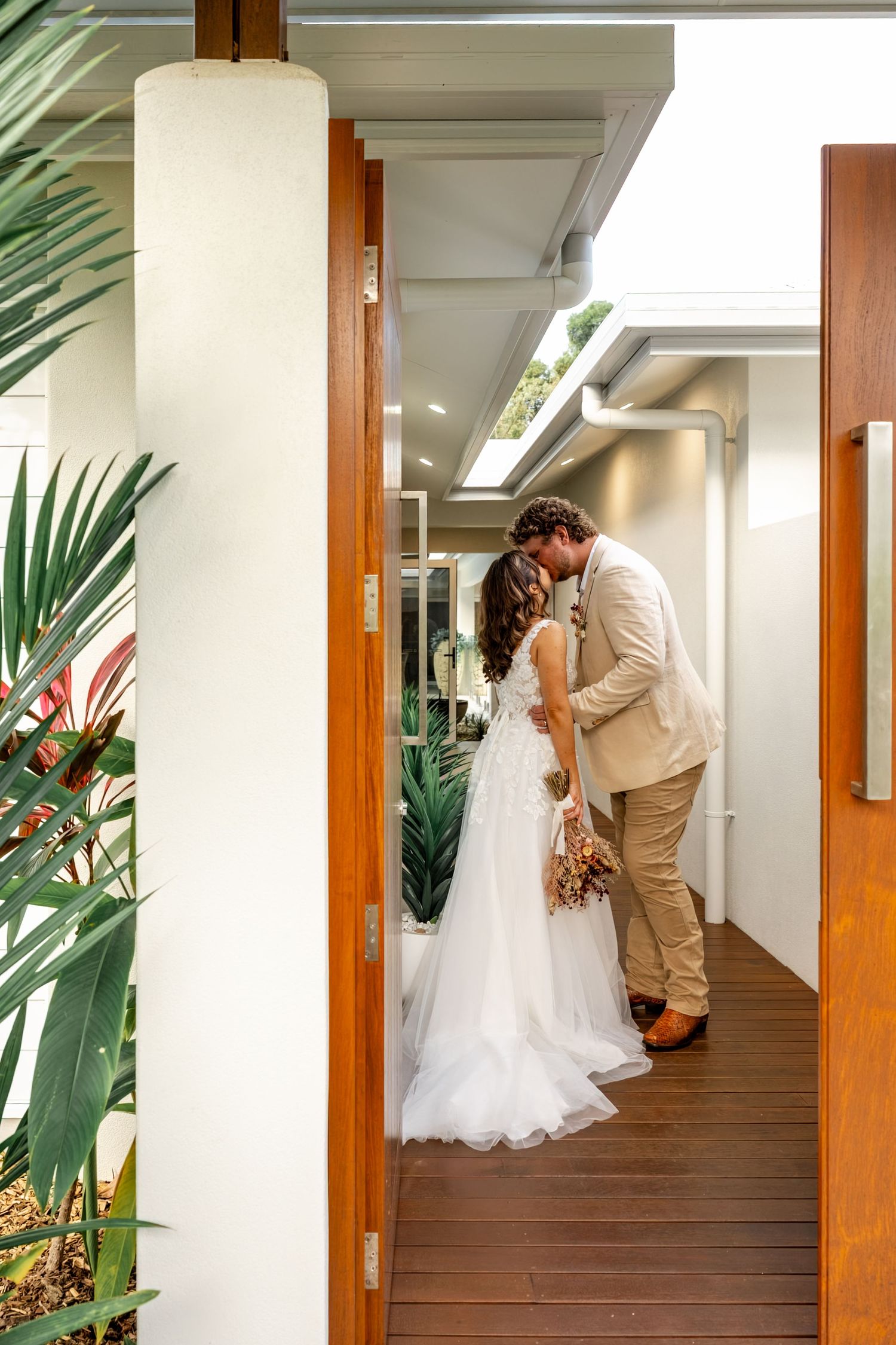 Bride and groom sharing a kiss in a modern timber hallway with tropical plants.