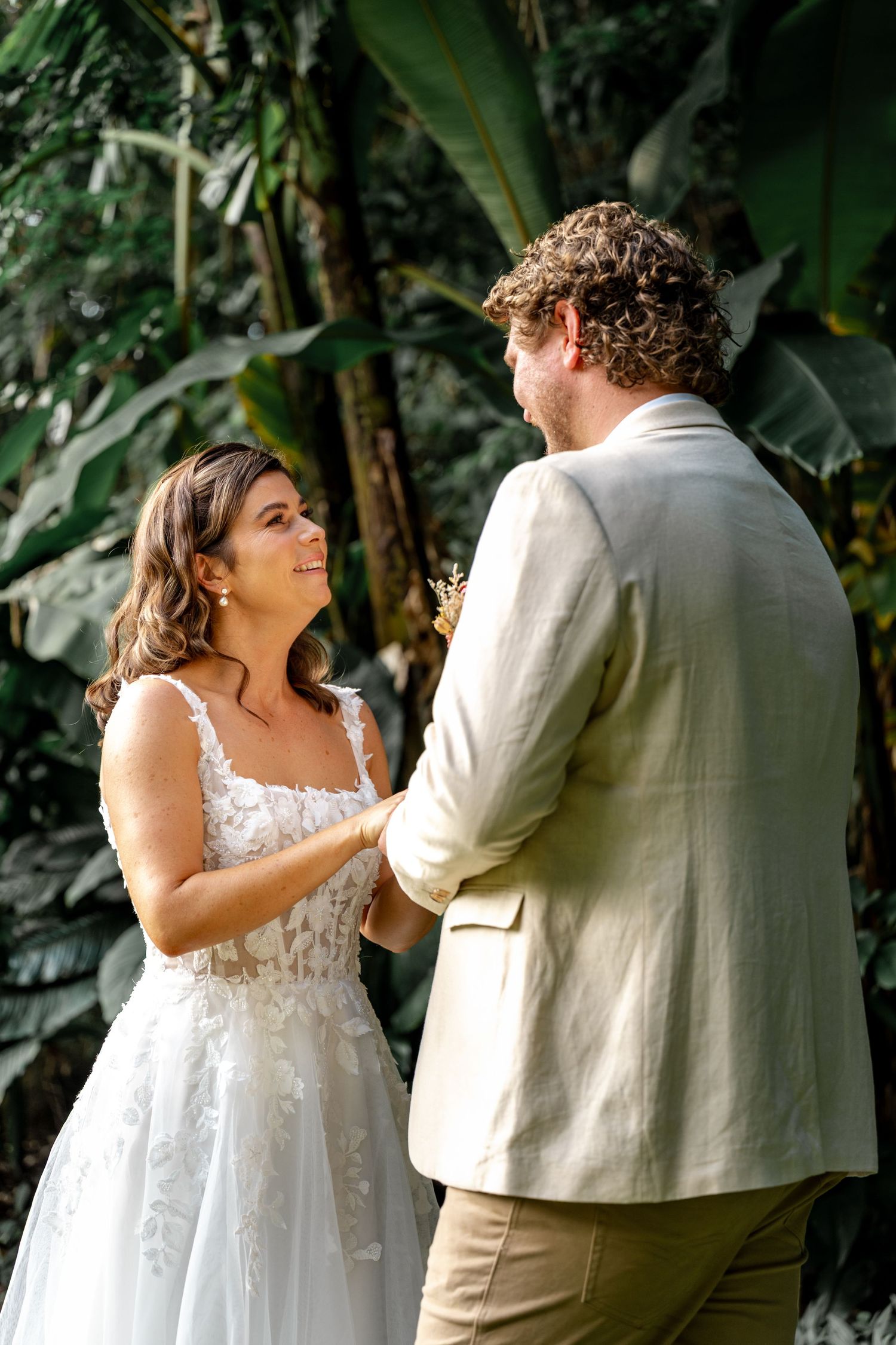 Bride smiles lovingly at groom while exchanging vows in lush tropical garden wedding ceremony.