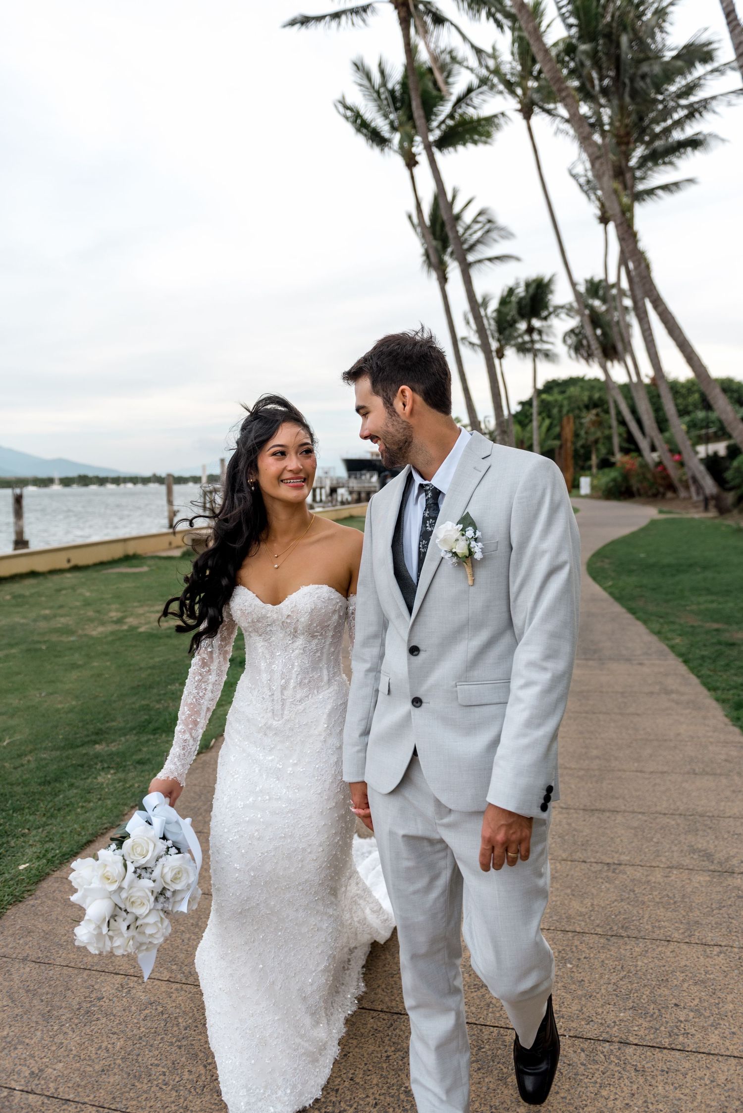 Bride and groom walk together smiling along a tropical palm tree-lined waterfront pathway.
