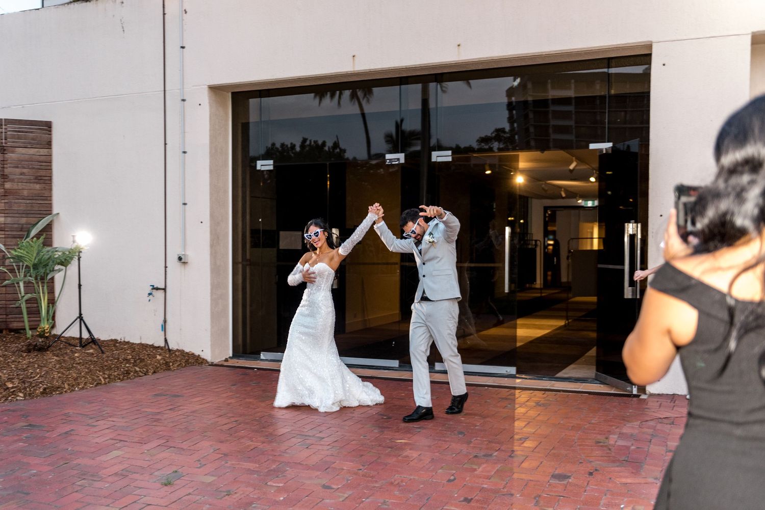 Bride and groom making grand entrance through glass doors as photographer captures the joyful moment outside venue.