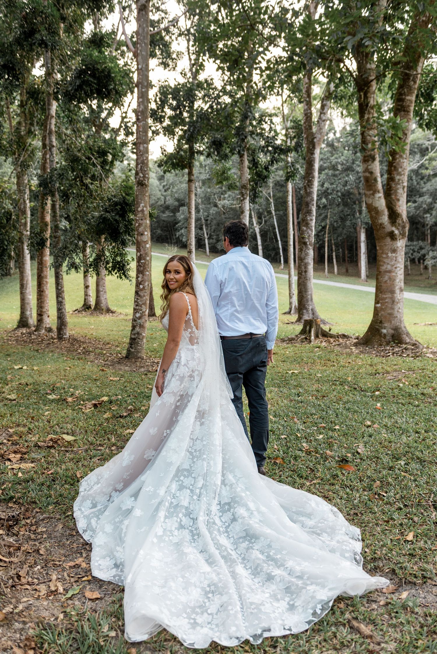 Bride in a lace gown with long train glances back while walking with groom through a grove of tall trees.