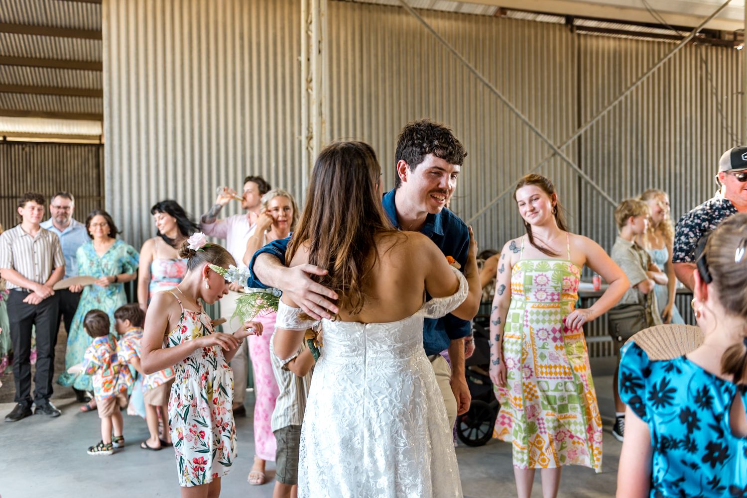 Bride and groom share first dance surrounded by smiling wedding guests inside a rustic barn venue.