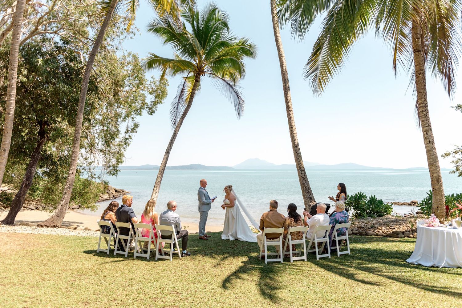 Wide shot of intimate tropical beach wedding ceremony with guests seated under palm trees.