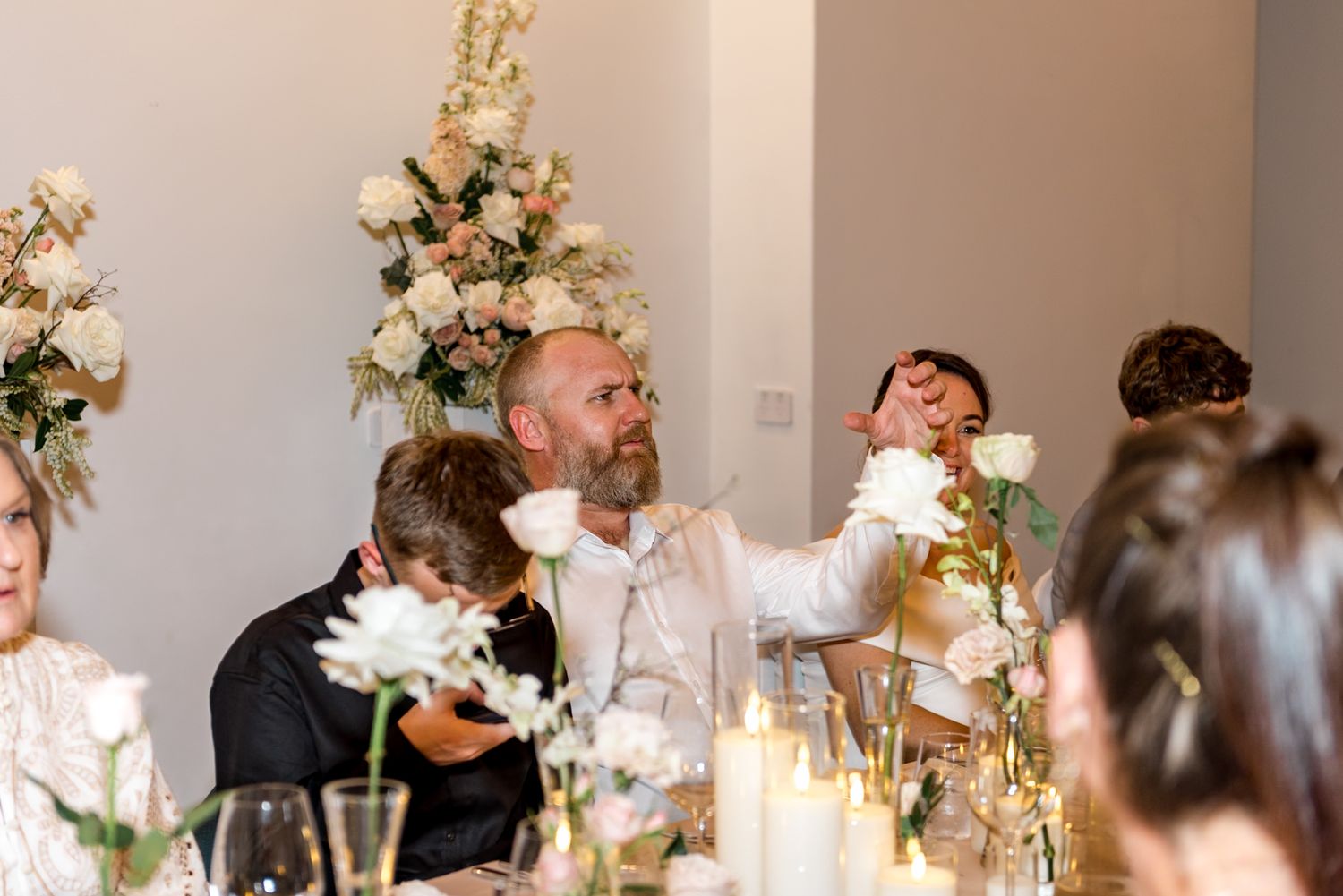 Wedding guest reaches across candlelit reception table decorated with white roses and floral arrangements.