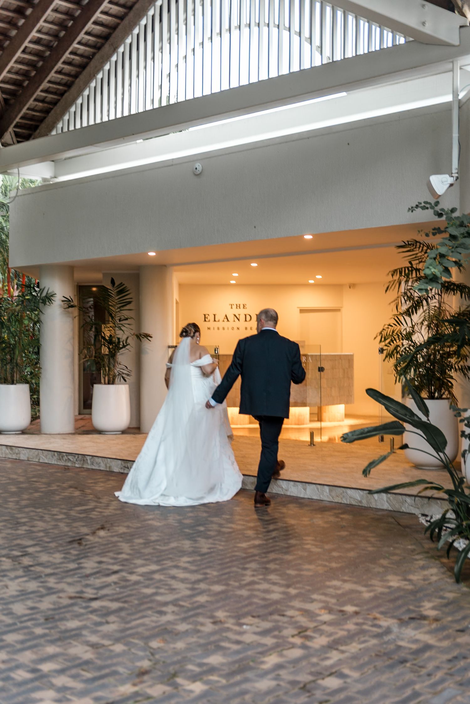 Bride in white gown and escort approaching The Elandra wedding venue entrance.