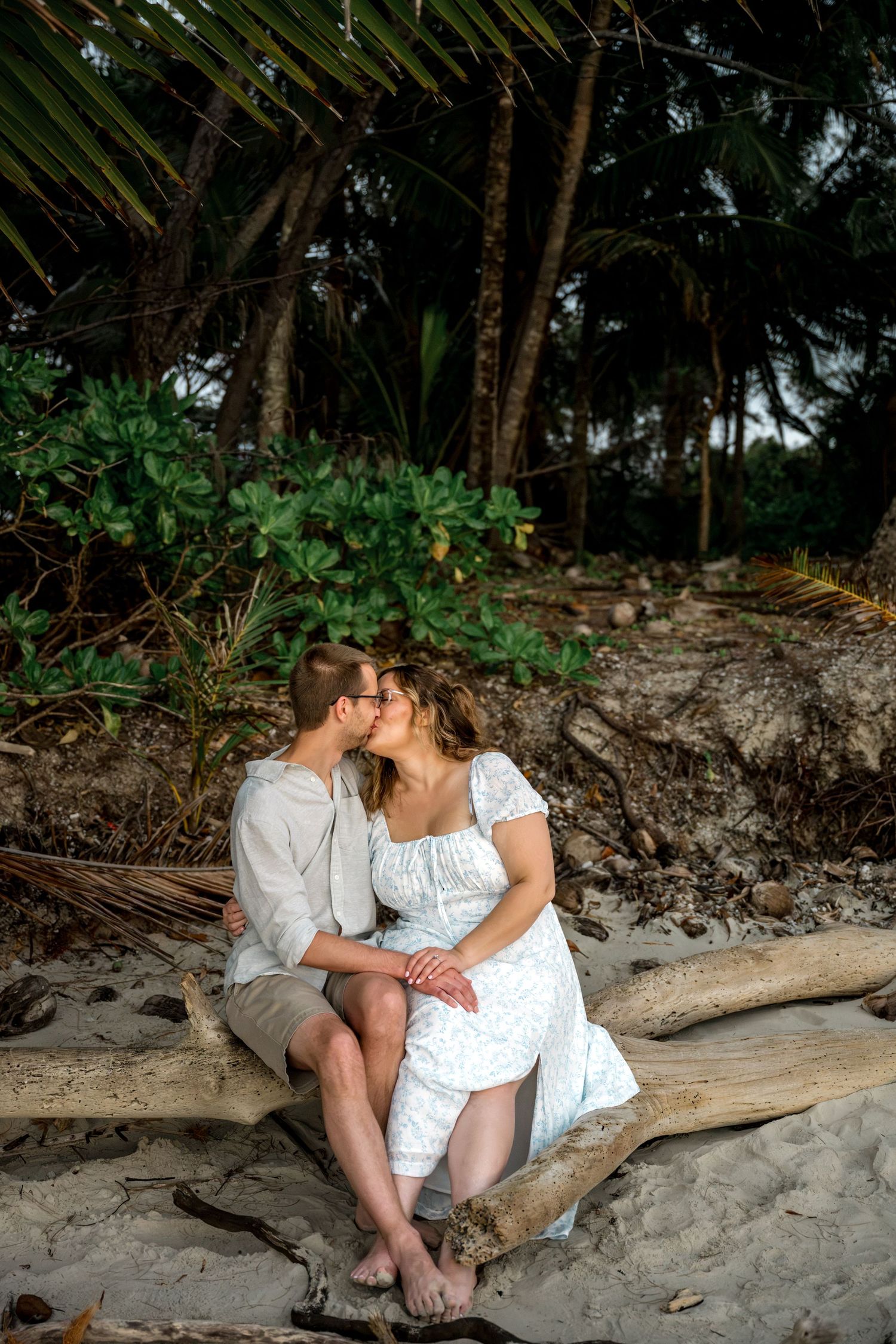 Engaged couple kissing intimately on driftwood log with palm trees and tropical foliage in background at Four Mile Beach, Port Douglas.