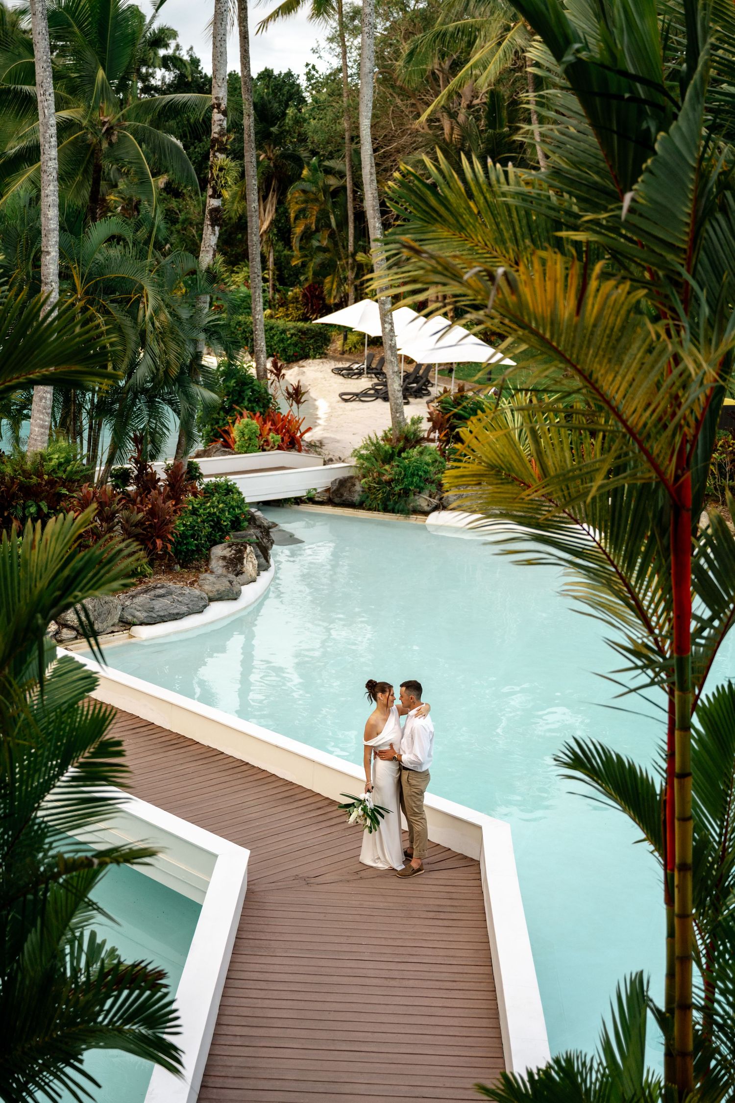 Aerial view of wedding couple standing on a resort deck surrounded by tropical palms and pool at Sheraton Grand Mirage Port Douglas.