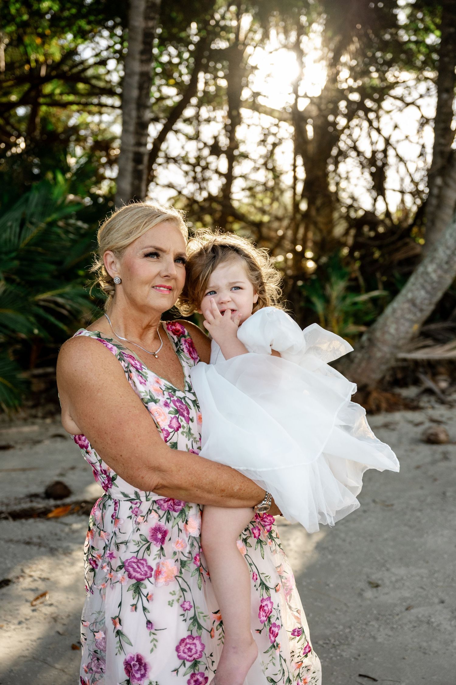 Grandmother in floral dress holding a young girl in white dress on a sunny beach at Four Mile Beach, Port Douglas.