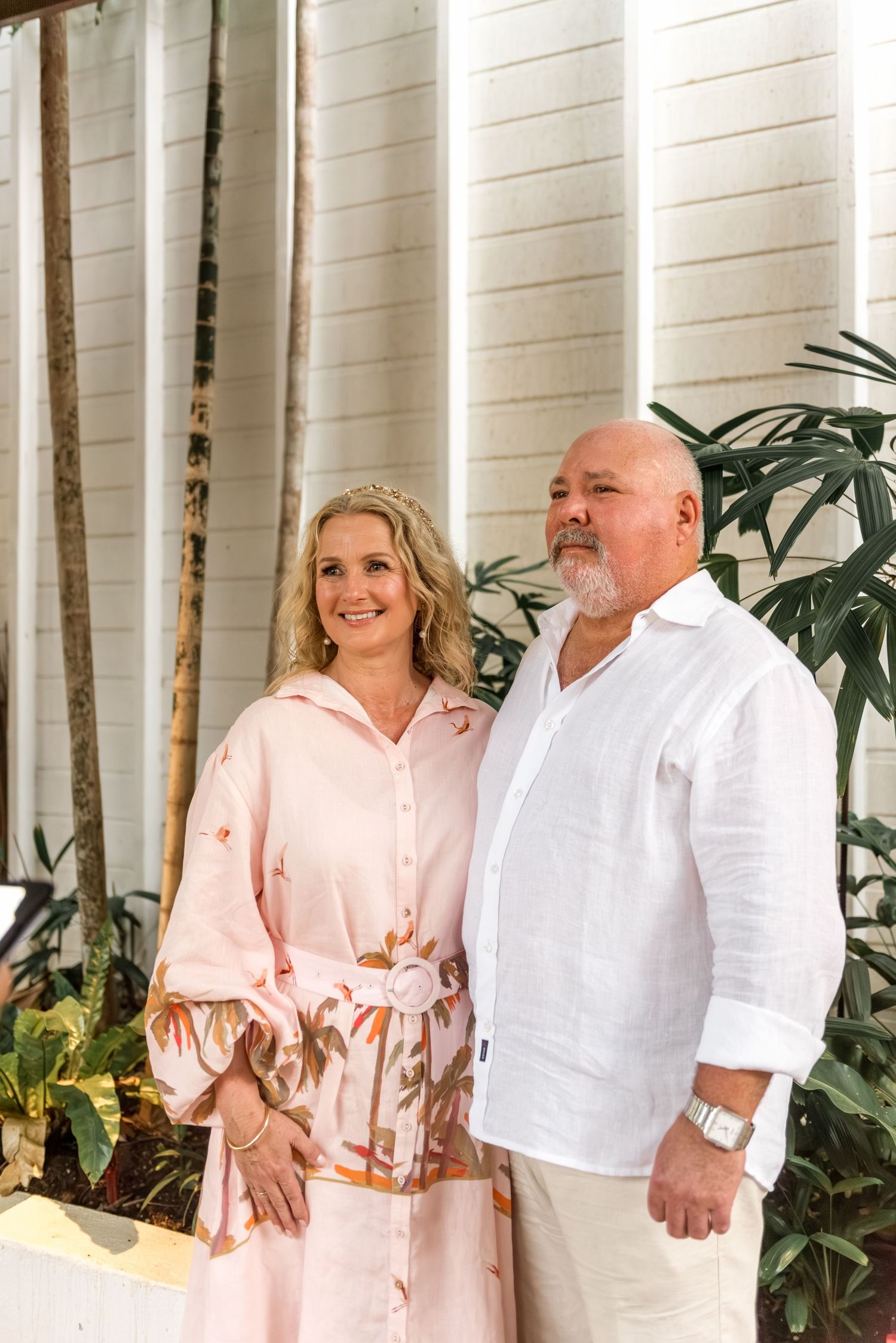 Elegant mature couple standing together in front of tropical plants and a white slatted wall.