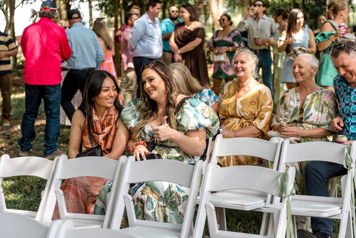 Wedding guests smiling and chatting while seated on white chairs at an outdoor ceremony.