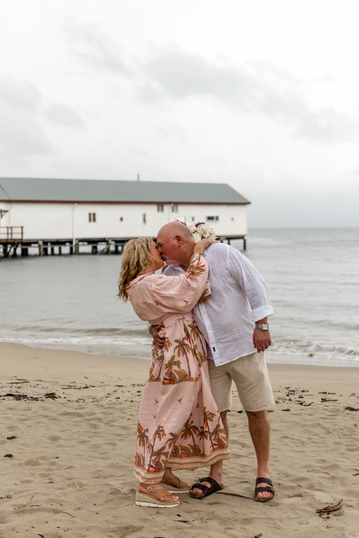 Couple sharing a passionate kiss on a sandy beach with a historic white boathouse behind them.