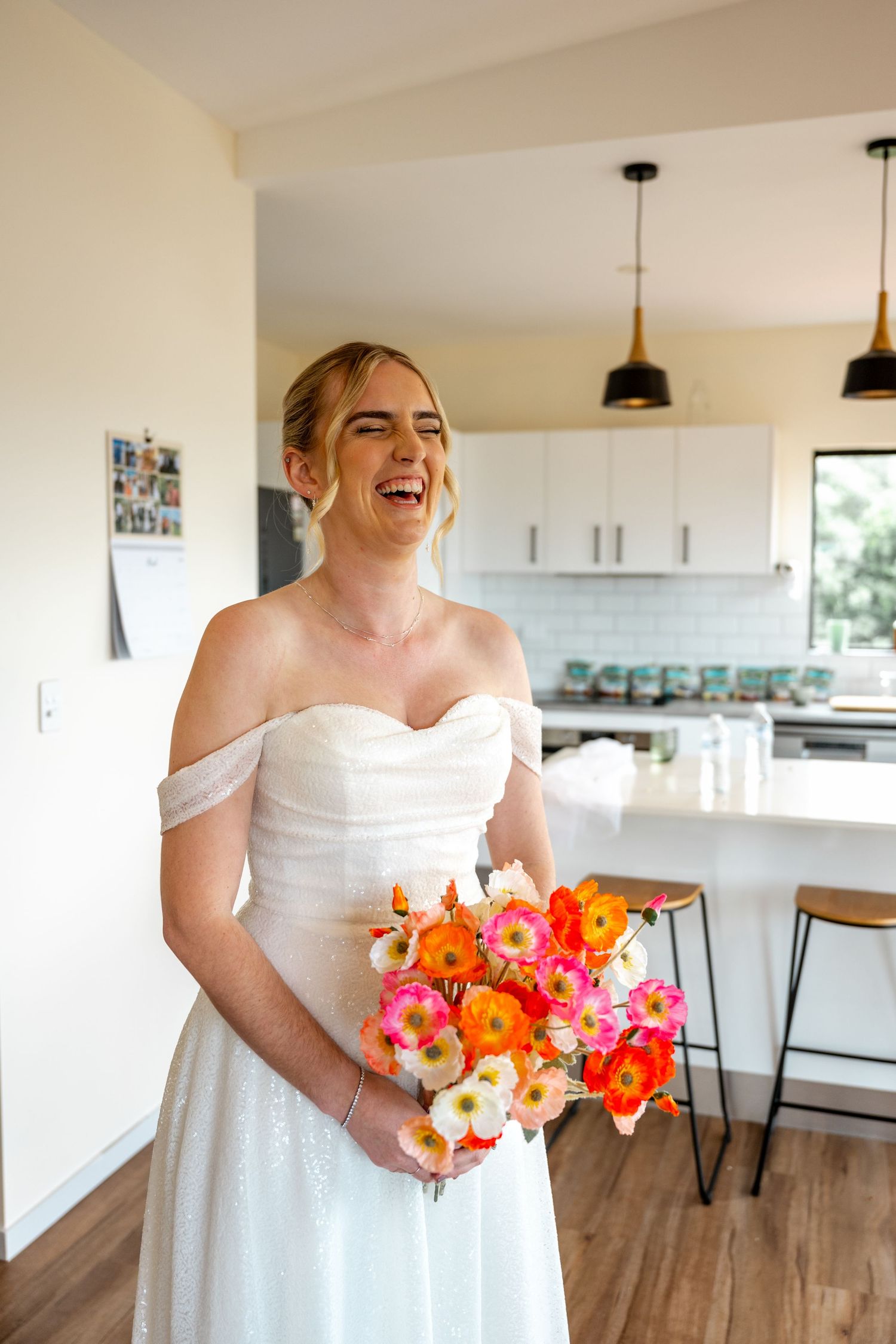 Laughing bride in off-shoulder white gown holding vibrant orange and pink flower bouquet indoors.